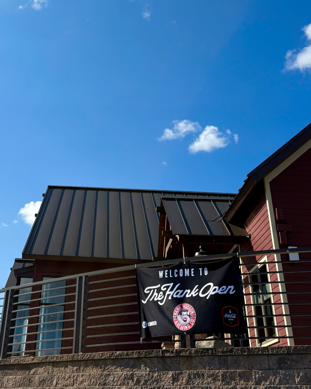 A black banner hanging on a balcony reads 'Welcome to The Hank Open' with logos for Hank Hudson Brewing Co., 84 Lumber, and Coca-Cola, with a clear blue sky and a building with a metal roof behind it.