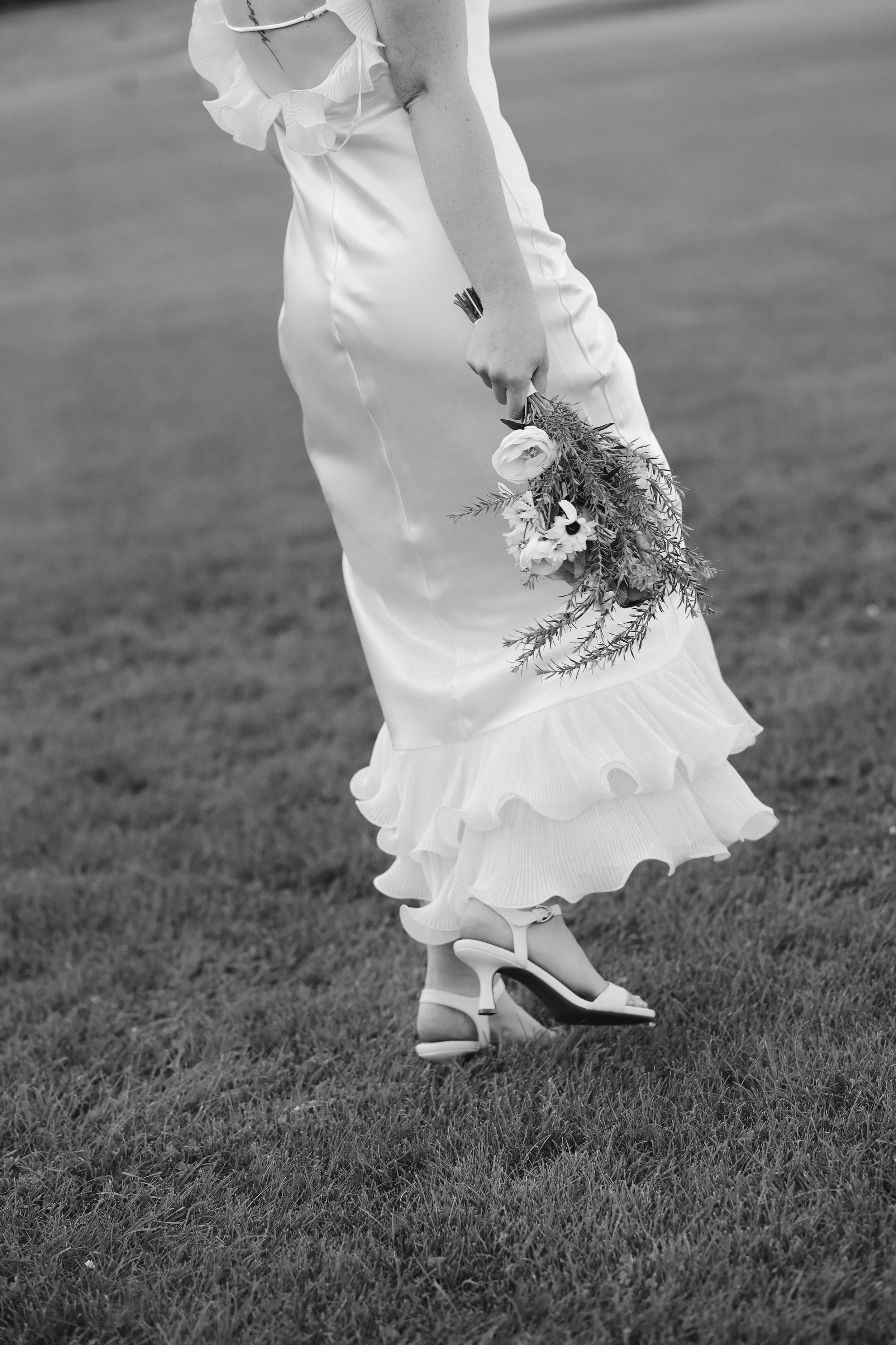 A person in a ruffled white dress holding a bouquet of flowers, wearing high heels, standing on grass with a blurred background.