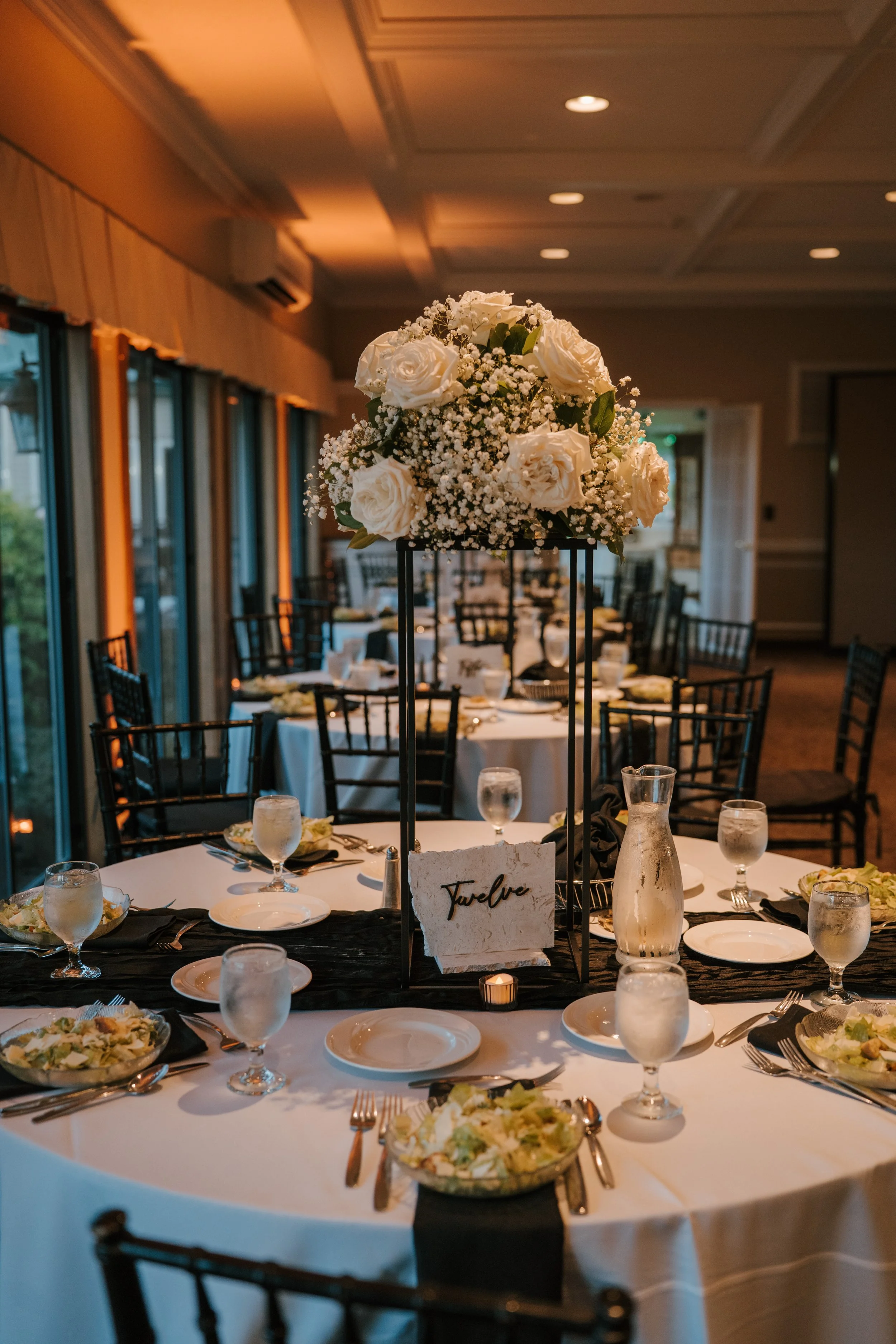 Elegant indoor wedding reception tables with floral centerpieces of white roses and baby's breath, set with place settings, glasses, and tableware, in a warmly lit room with windows and high ceiling.