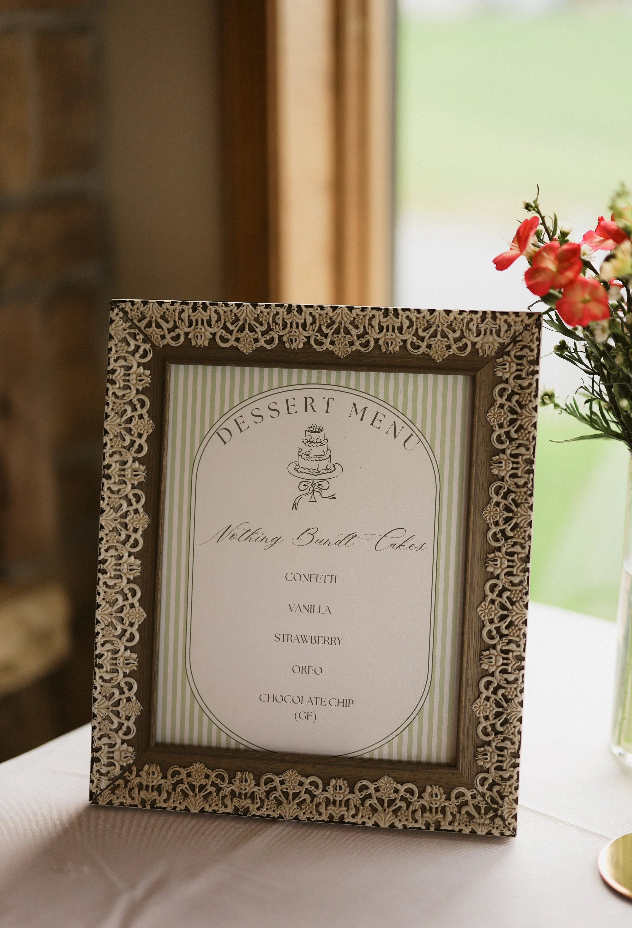 A framed dessert menu with a decorative lace-like border, listing various sweet treats including confetti, vanilla, strawberry, Oreo, chocolate chip, and gluten-free options, placed on a white tablecloth next to a vase of pink and white flowers.