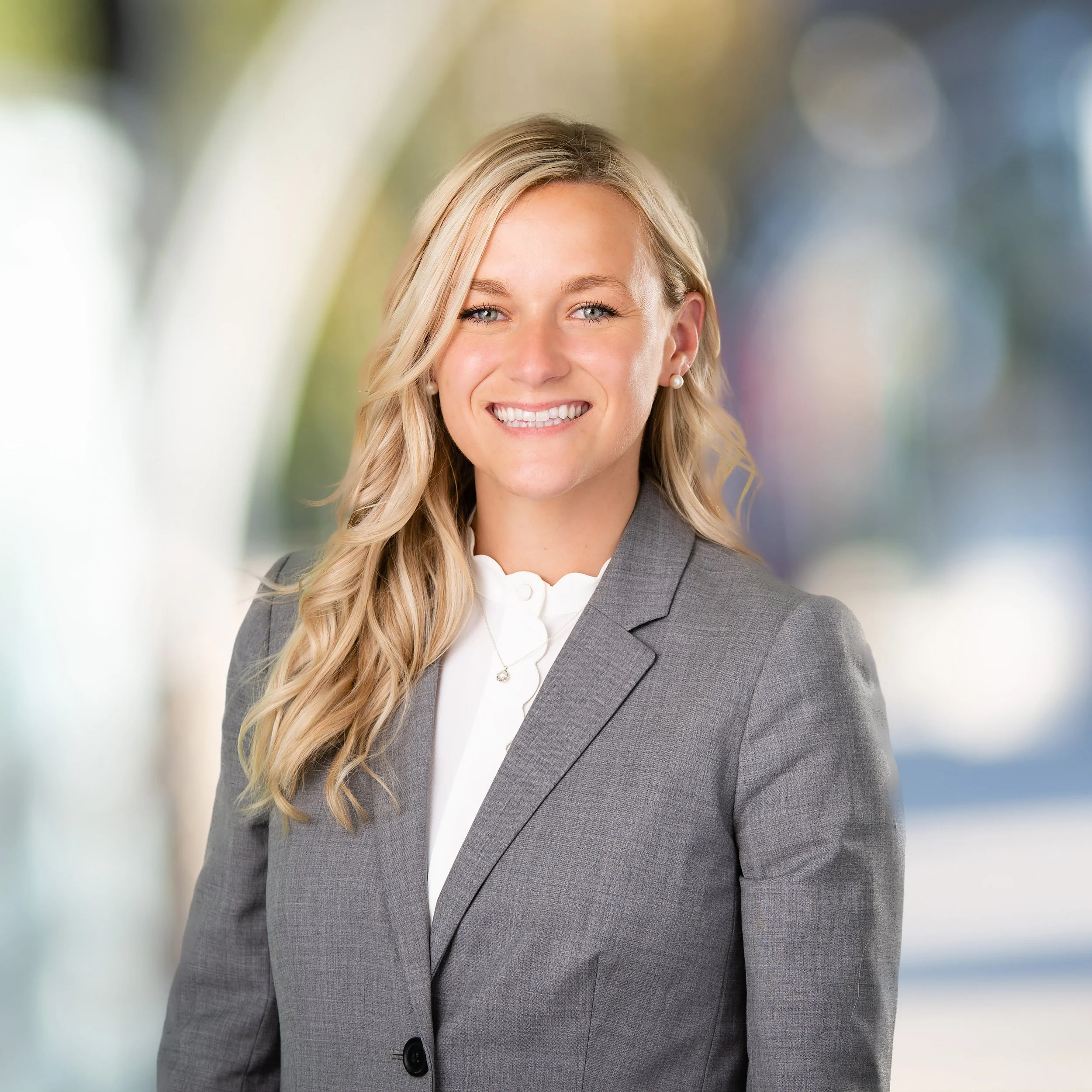 Professional woman with blonde hair in a gray suit and white blouse, smiling against a blurred outdoor background.