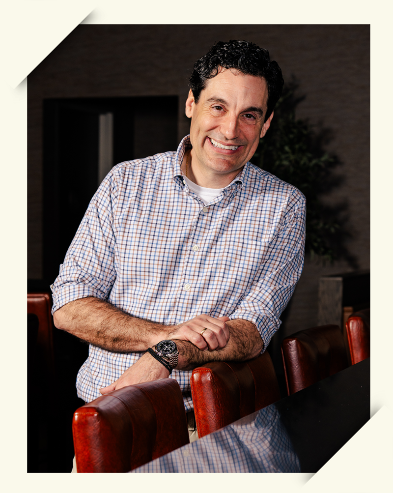 A smiling man with dark curly hair, wearing a checkered shirt and a watch, leaning on a conference table in a modern office setting.