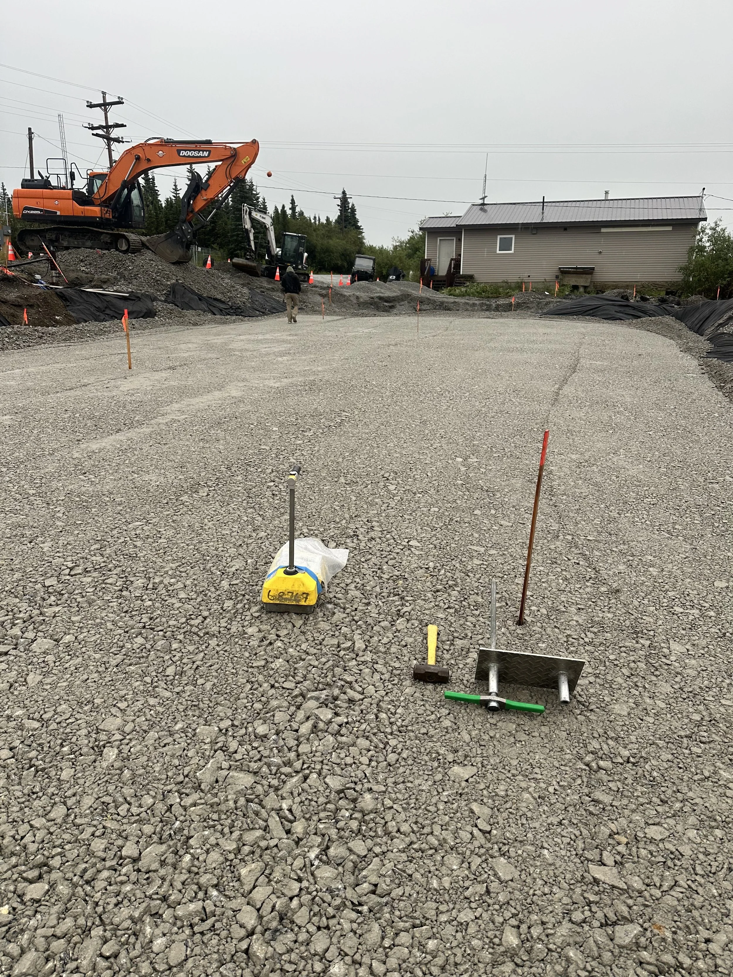 A construction site with gravel ground, orange excavator, black machinery, and a person walking. There is a building in the background and orange safety cones.