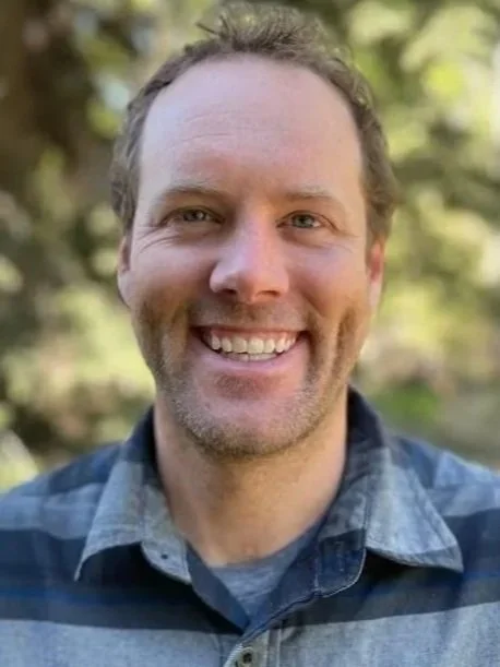 A man with light brown hair, a beard, and a smiling face, wearing a plaid shirt outdoors with trees in the background.