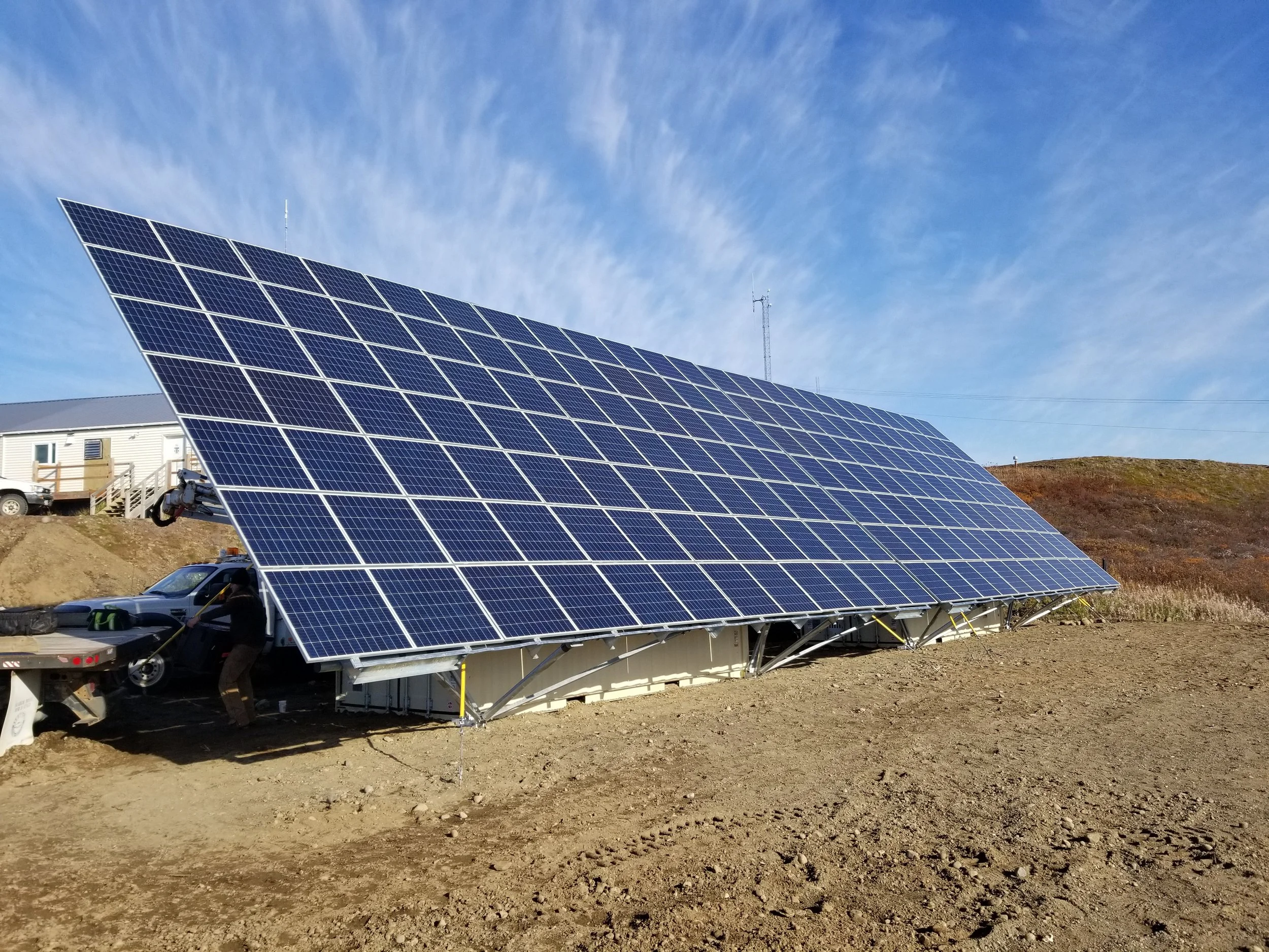 A solar panel installation on a dirt field with a building, a vehicle, and a person working nearby, under a blue sky with wispy clouds.