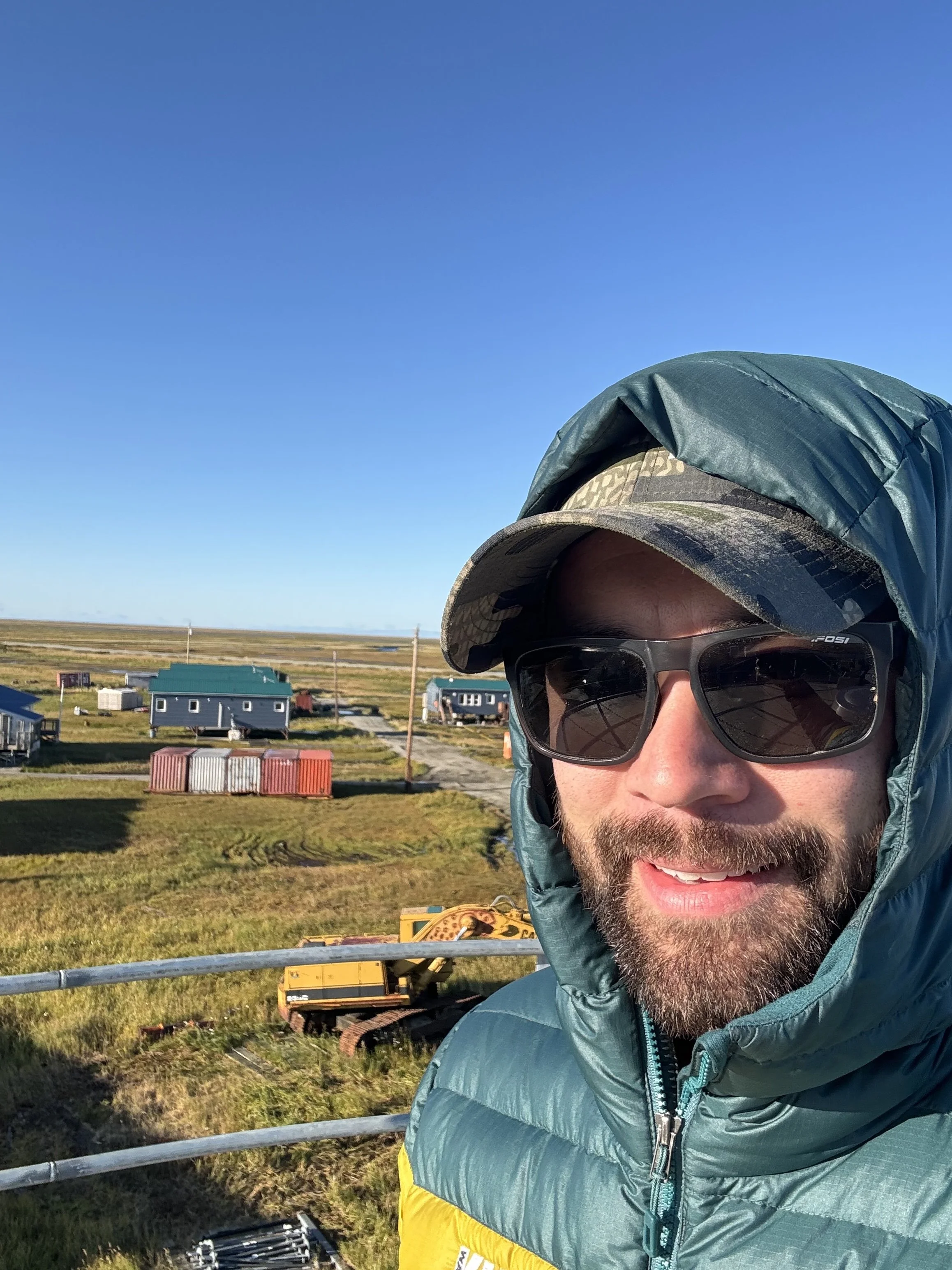 A man outdoors in a green jacket and camo hat, wearing sunglasses, smiling at the camera against a clear blue sky with buildings, fields, and construction equipment in the background.