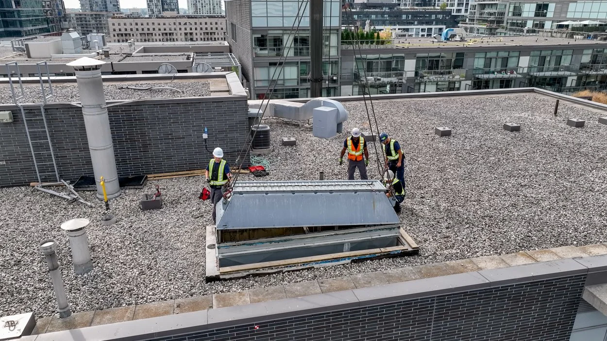 Four male construction workers in safety equipment install an HVAC system on the roof of a tall building. A crane lowers the HVAC system into place.
