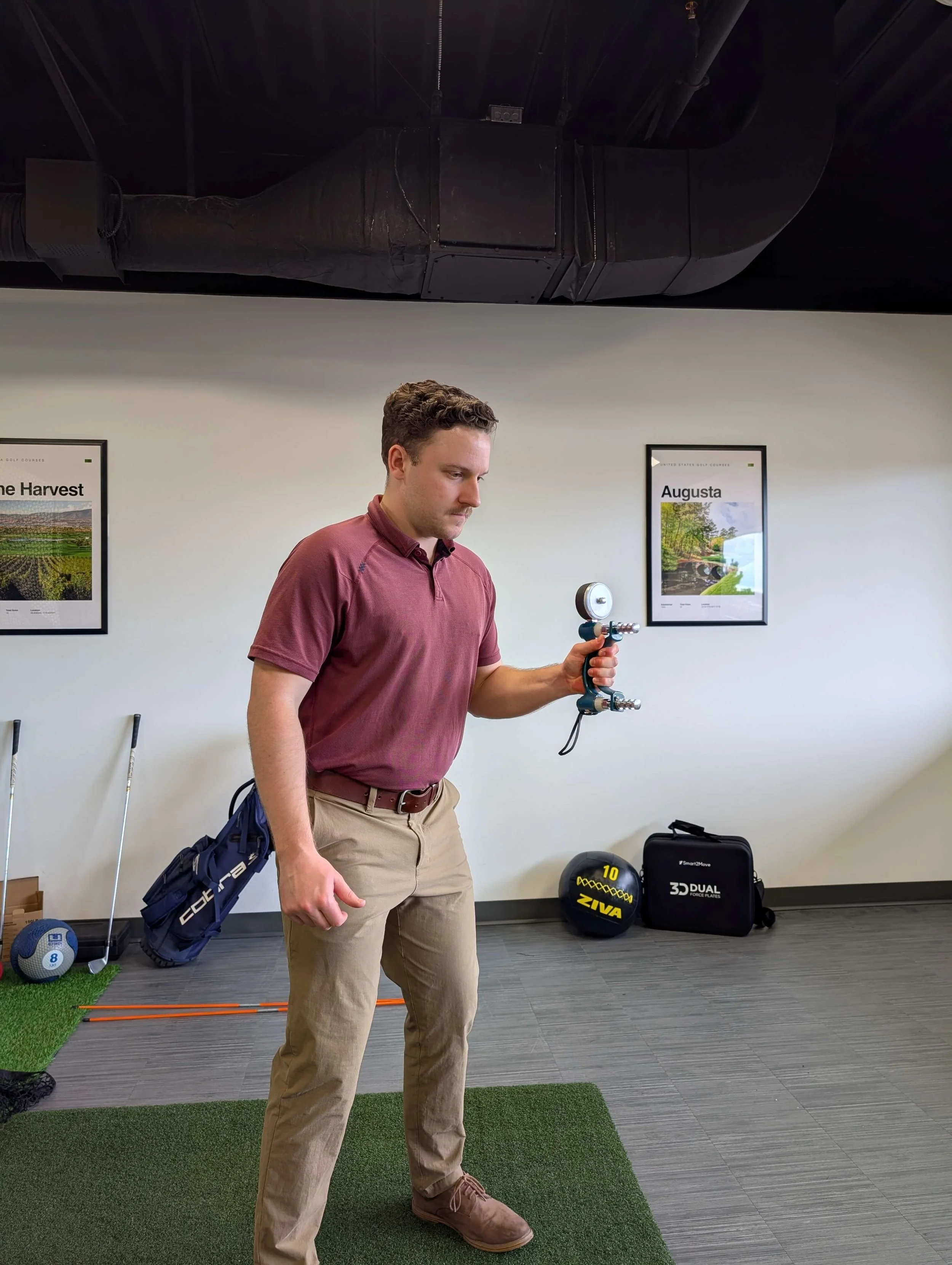 A man in a maroon polo shirt and khaki pants holding a golf swing trainer in a room with golf equipment, framed pictures on the wall, and a small artificial grass mat on the floor to improve his golf game using golf instruction.