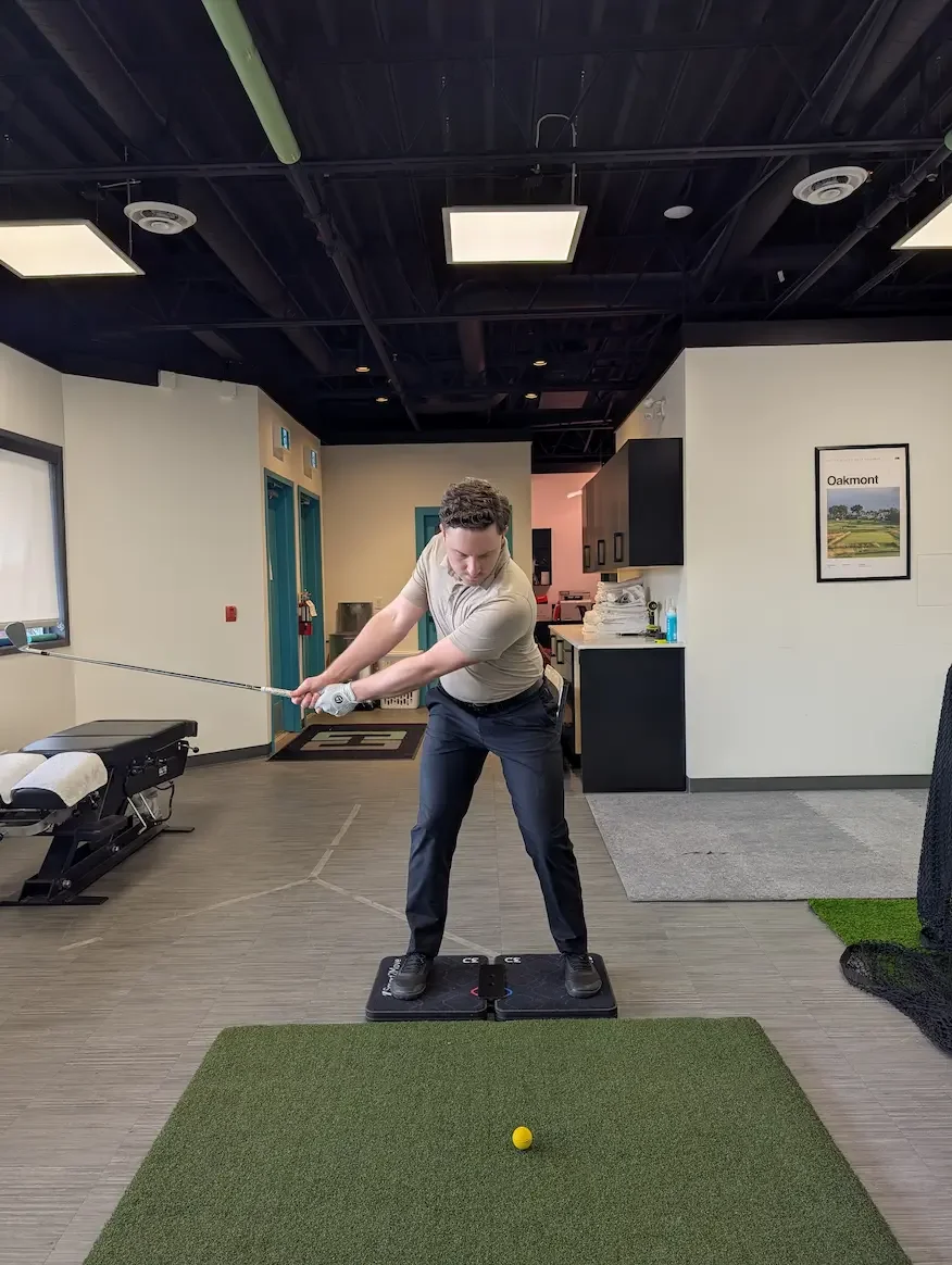 Man practicing golf swing indoors on a swing trainer mat with a yellow golf ball on a green mat