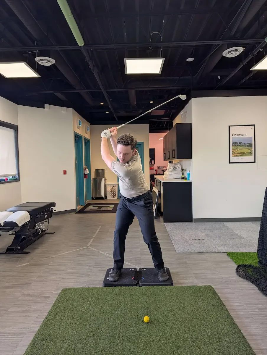 A man practicing golf swings indoors, standing on a black mat on a small green artificial turf, with a yellow golf ball in front of him.