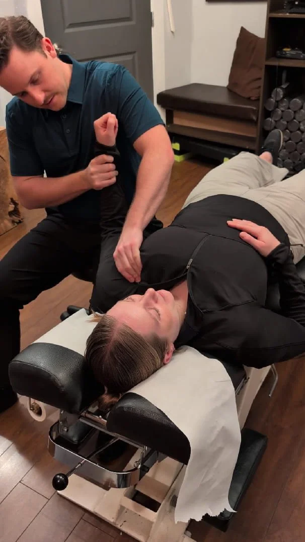 A woman lying face up on a chiropractic adjusting table, with a hand on her chest and eyes closed, while a man in a blue shirt and black pants holds her arm and appears to be performing an adjustment in a treatment room.