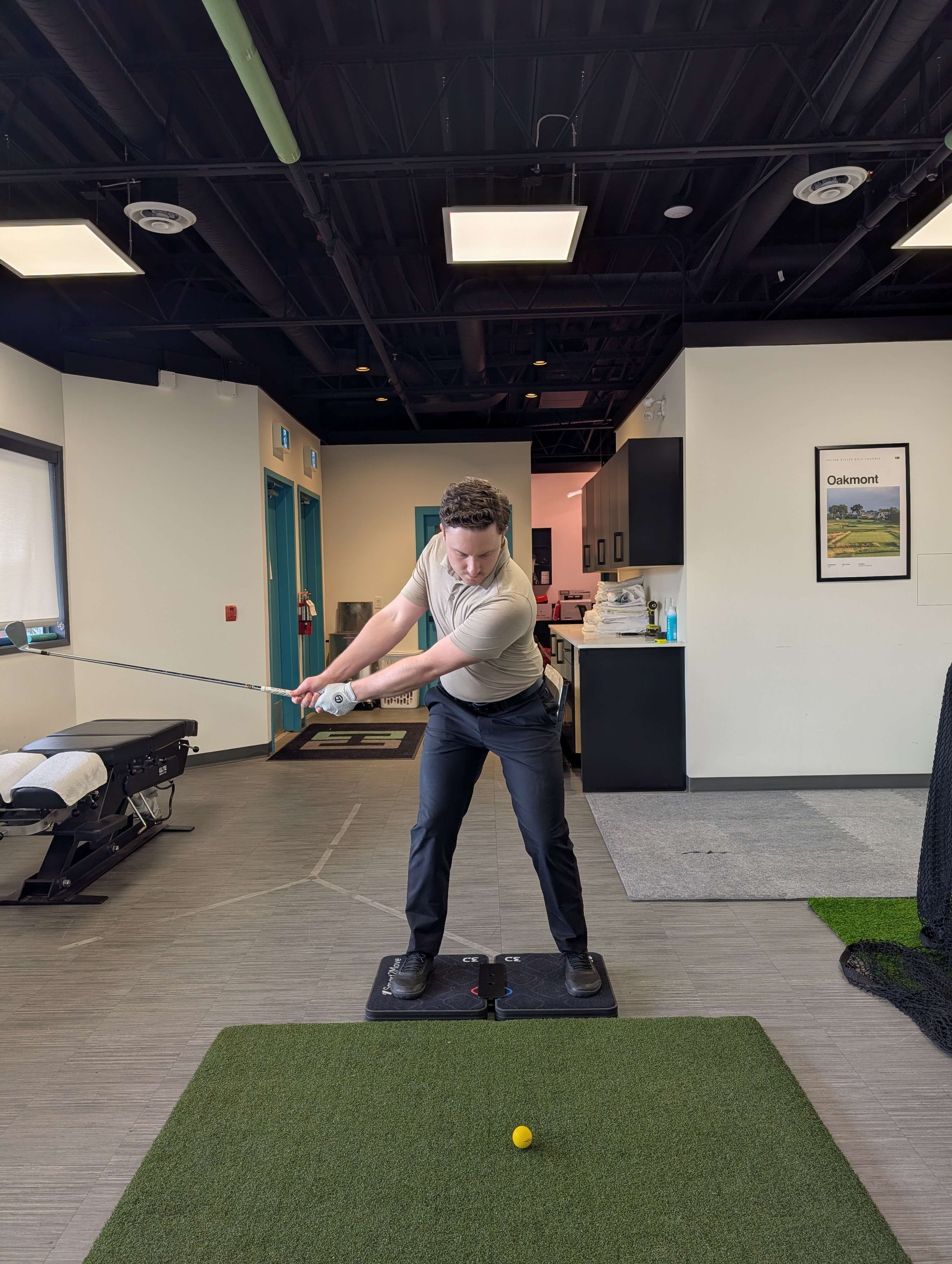 A man practicing during a golf lesson indoors, preparing to hit a golf ball with a club, standing on a mat in a room that appears to be a golf simulation or training space.
