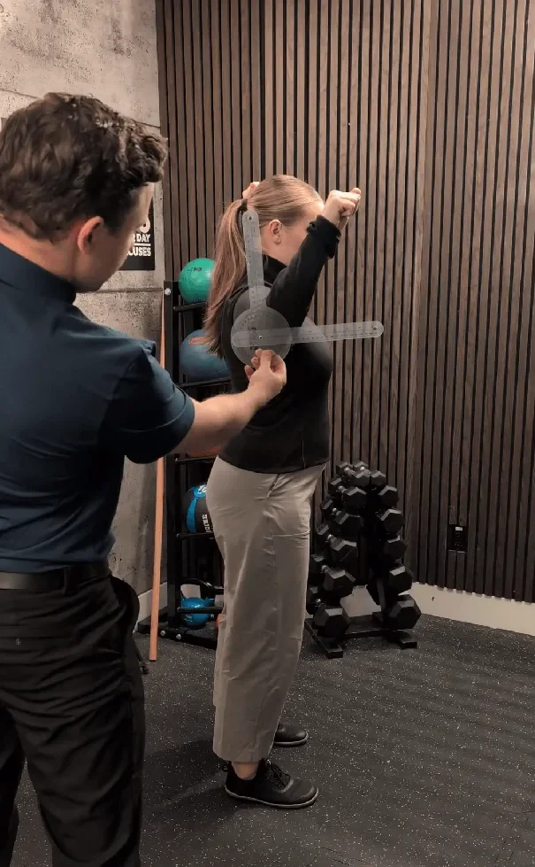 A woman is performing an exercise with a measurement device held by a trainer in a gym. The woman is standing with her arm lifted, and the trainer is taking her measurements.