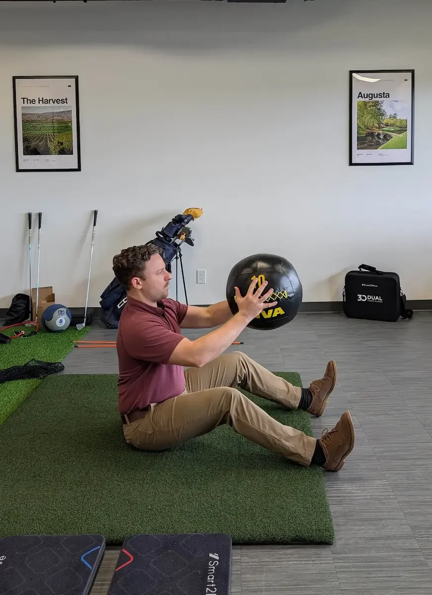 A man sitting on a green mat in an indoor space, holding a black medicine ball with yellow writing, with gym equipment and framed posters on the wall behind him.