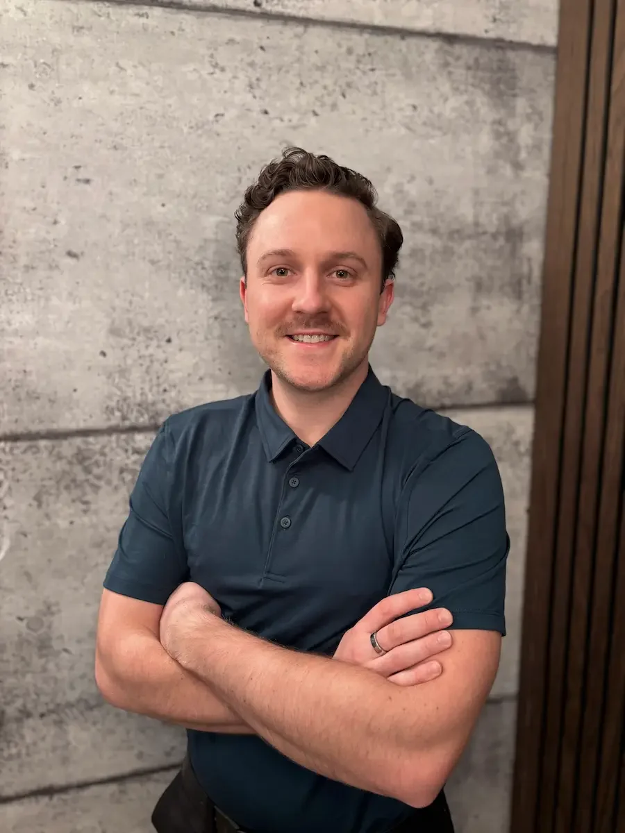 A man with short, curly brown hair and a beard, smiling with arms crossed, wearing a dark blue polo shirt, standing against a concrete wall with a wooden panel on the right side.