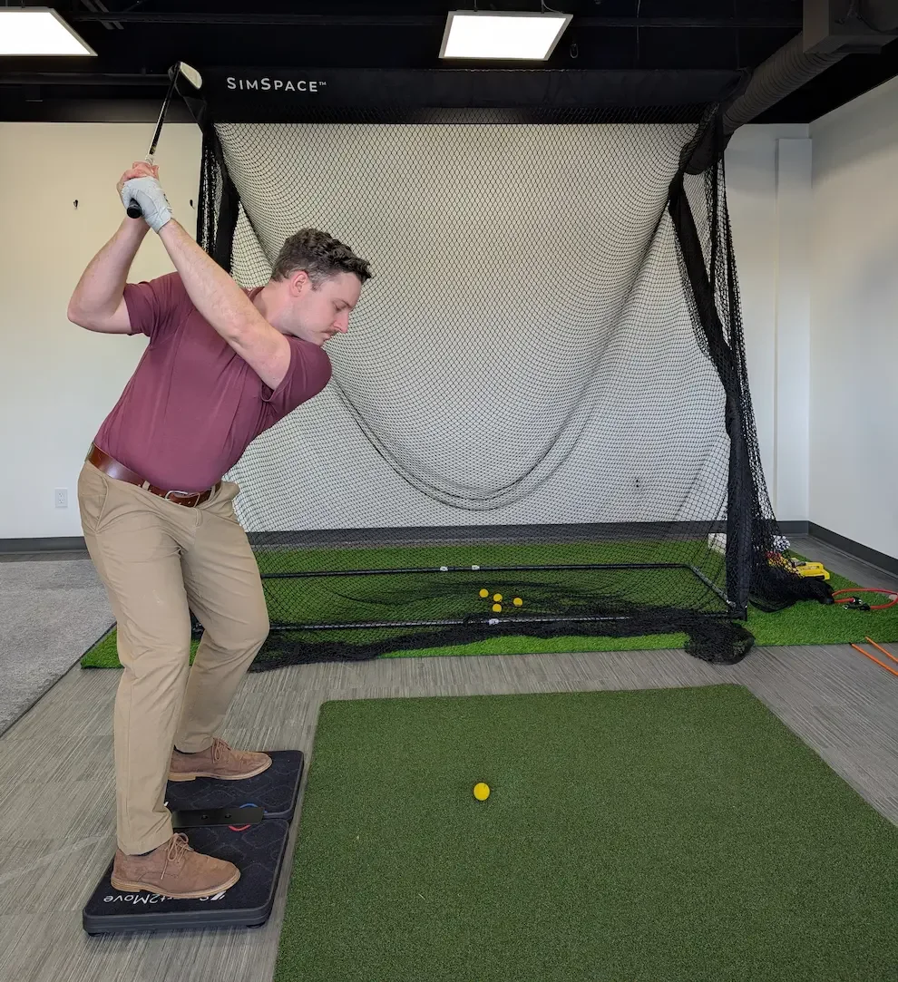 A man practicing golf indoors, swinging a golf club from a mat, with a golf net behind him for practicing swings, and golf balls scattered on the mat.