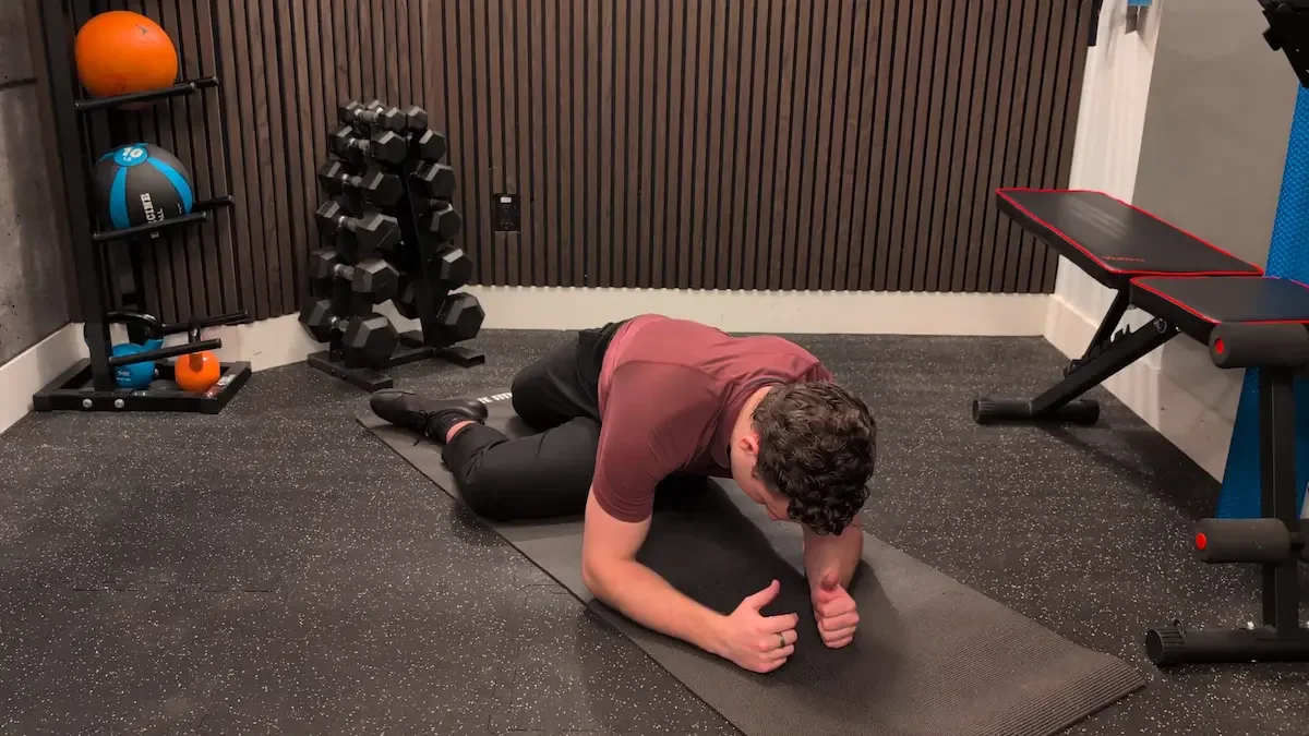 A man performing a forearm plank exercise on a black exercise mat in a home gym, with dumbbells, medicine balls, and workout equipment in the background.