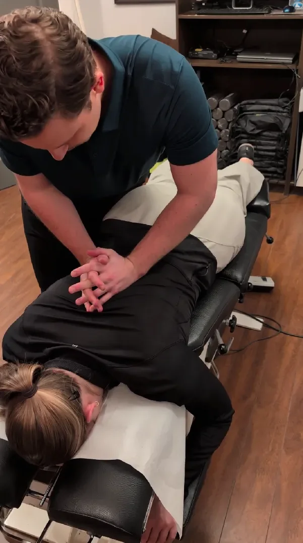 A chiropractor or massage therapist working on a patient lying face down on a treatment table in a room with wooden floors and shelves