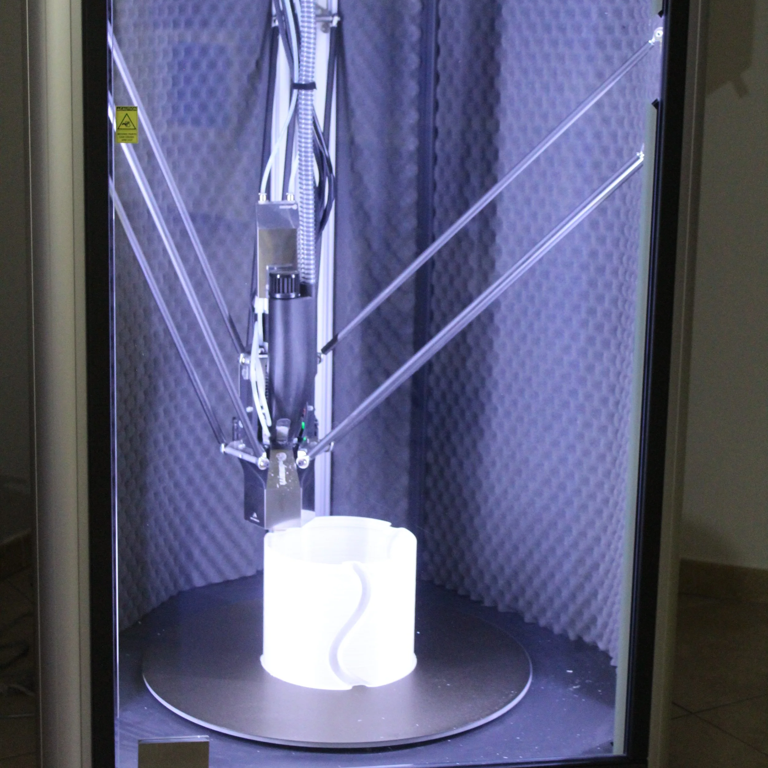 Immagine di una stampante 3D in funzione all’interno di una cabina insonorizzata, che sta stampando un oggetto bianco.