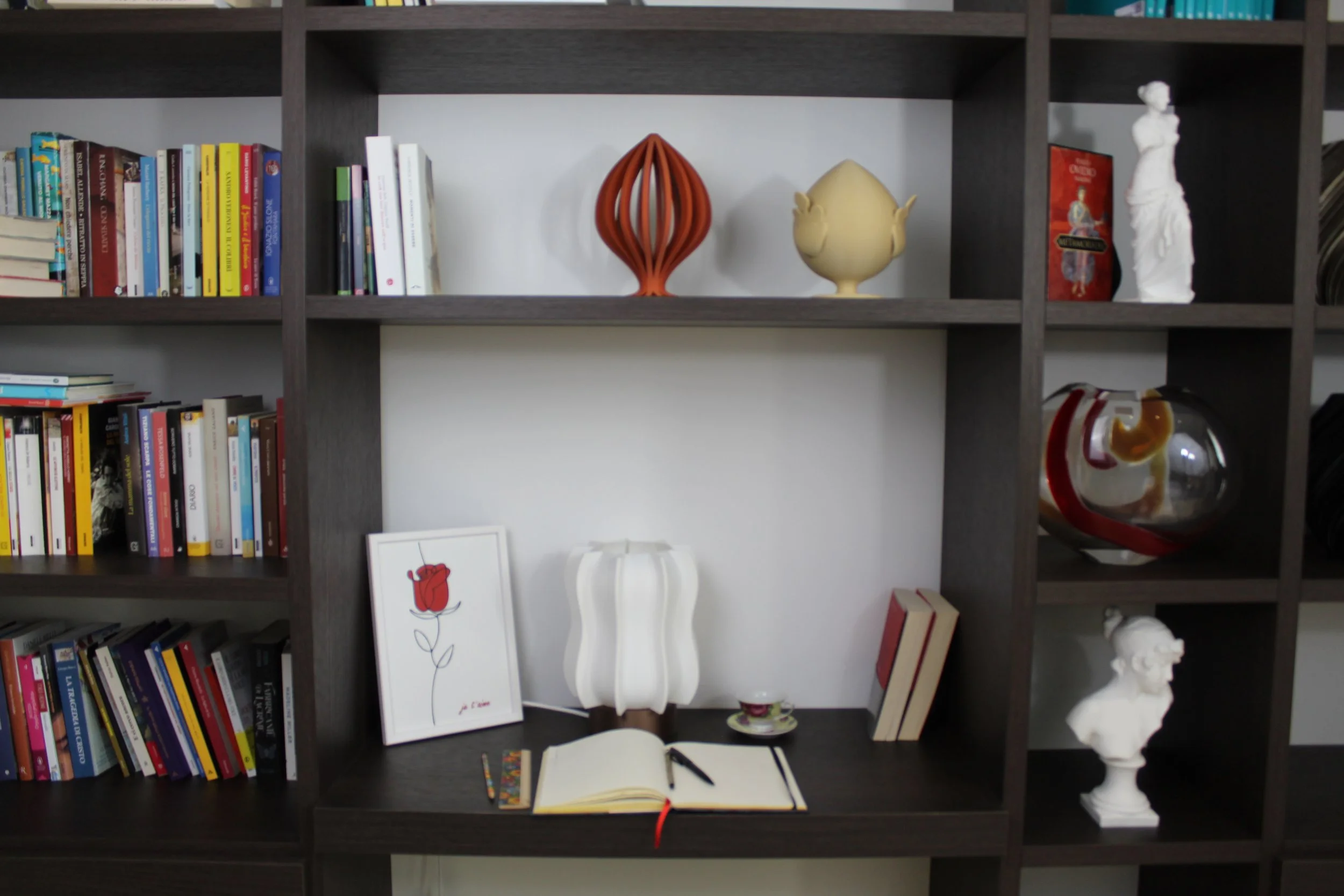 Scaffale con libri, decorazioni, e oggetti d'arte, tra cui vasi, statue e una lampada, affiancato da una scrivania con un quaderno aperto, una penna, una tazza e un disegno di un fiore rosso.