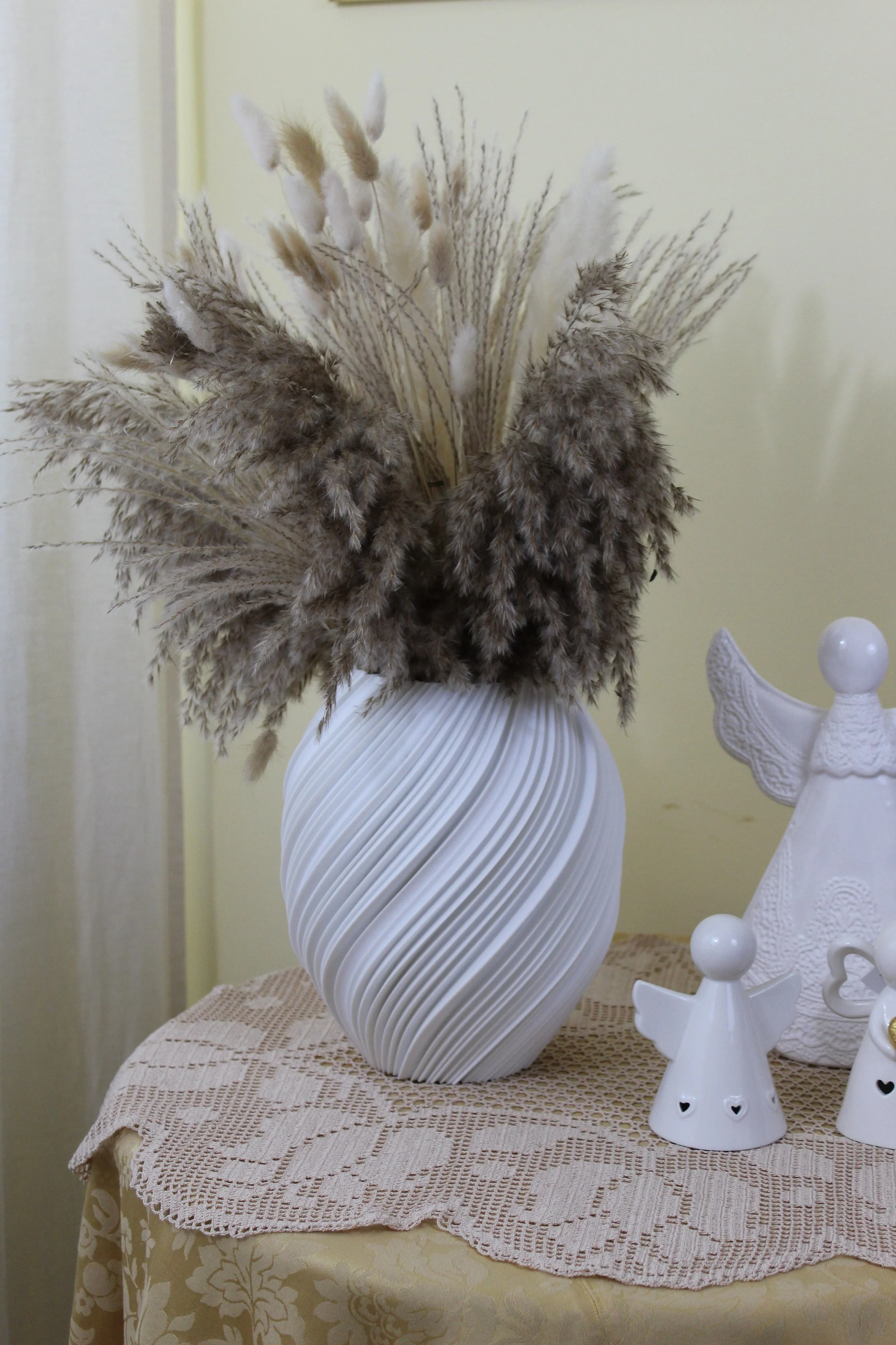 Vasetto bianco con decorazioni ondulate, contenente un mazzolino di pampas grass e altre piante secche di colore beige, bianco e grigio, su un tavolo con tovaglia ricamata