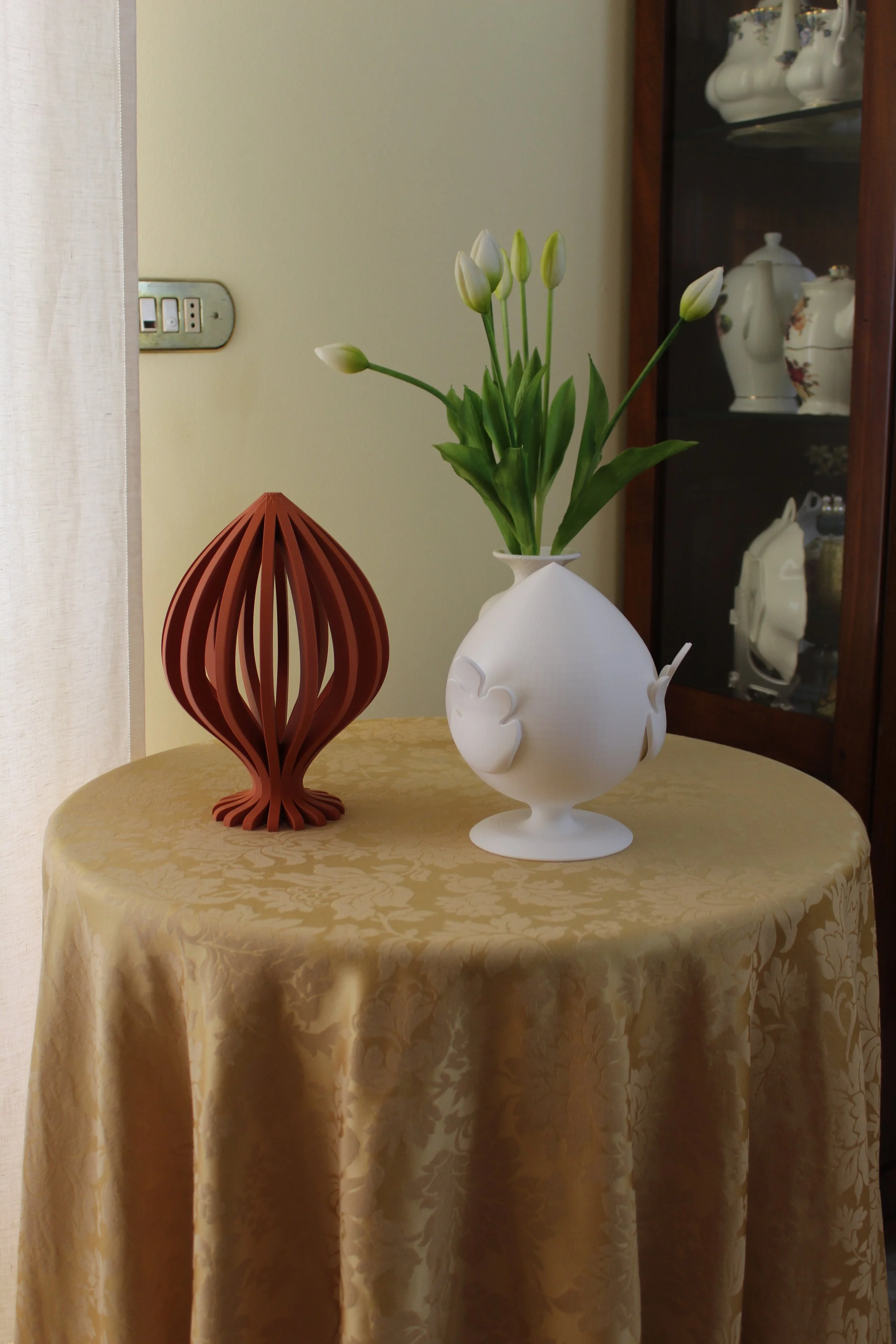 Vaso bianco con fiori di tulipano e decorazioni di foglie, a fianco una lanterna decorativa di legno, entrambi su un tavolo rotondo con tovaglia dorata.