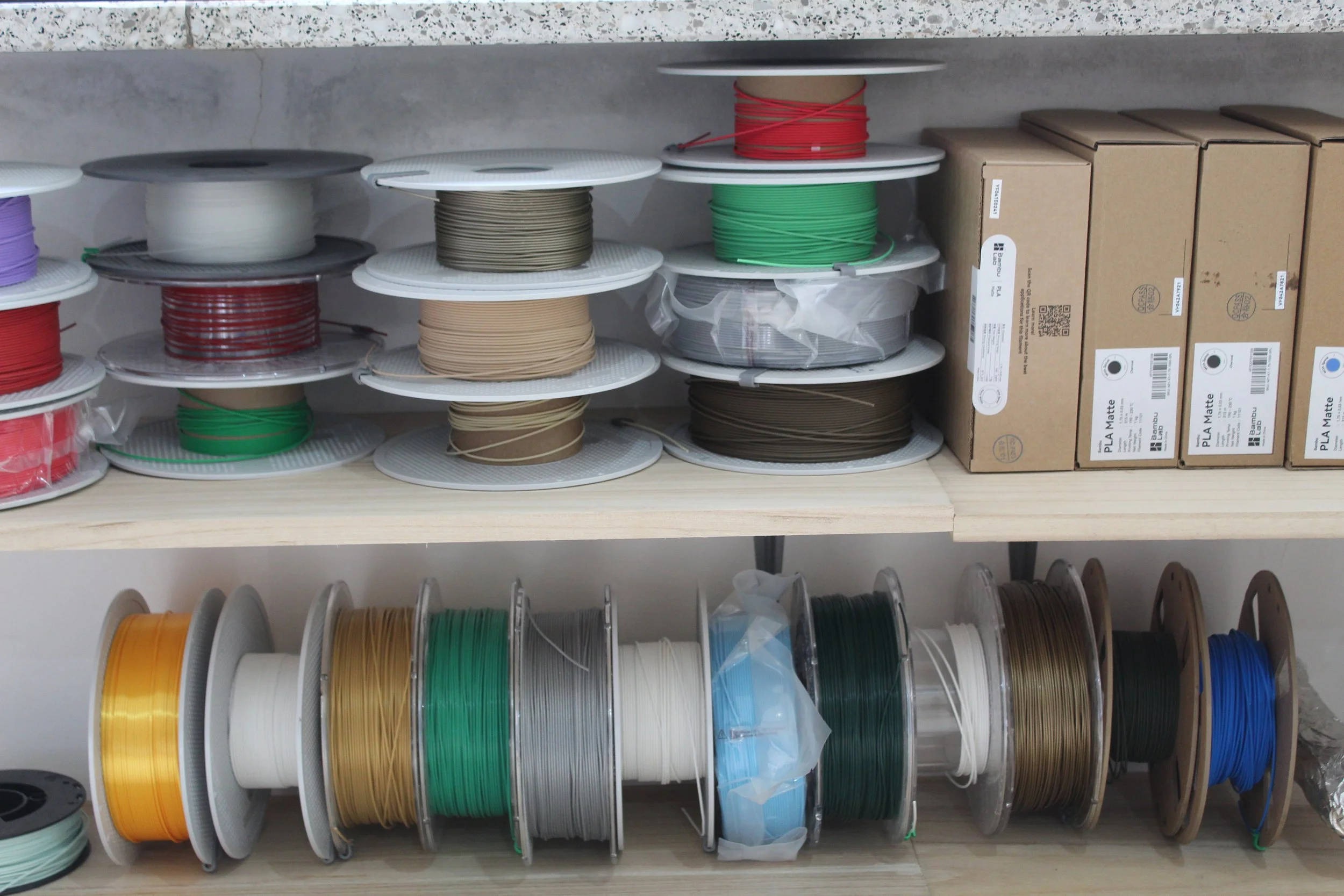 Spirali di filamenti di plastica di vari colori su un supporto in plastica, organizzati su ripiani di legno in un laboratorio o negozio di stampa 3D.