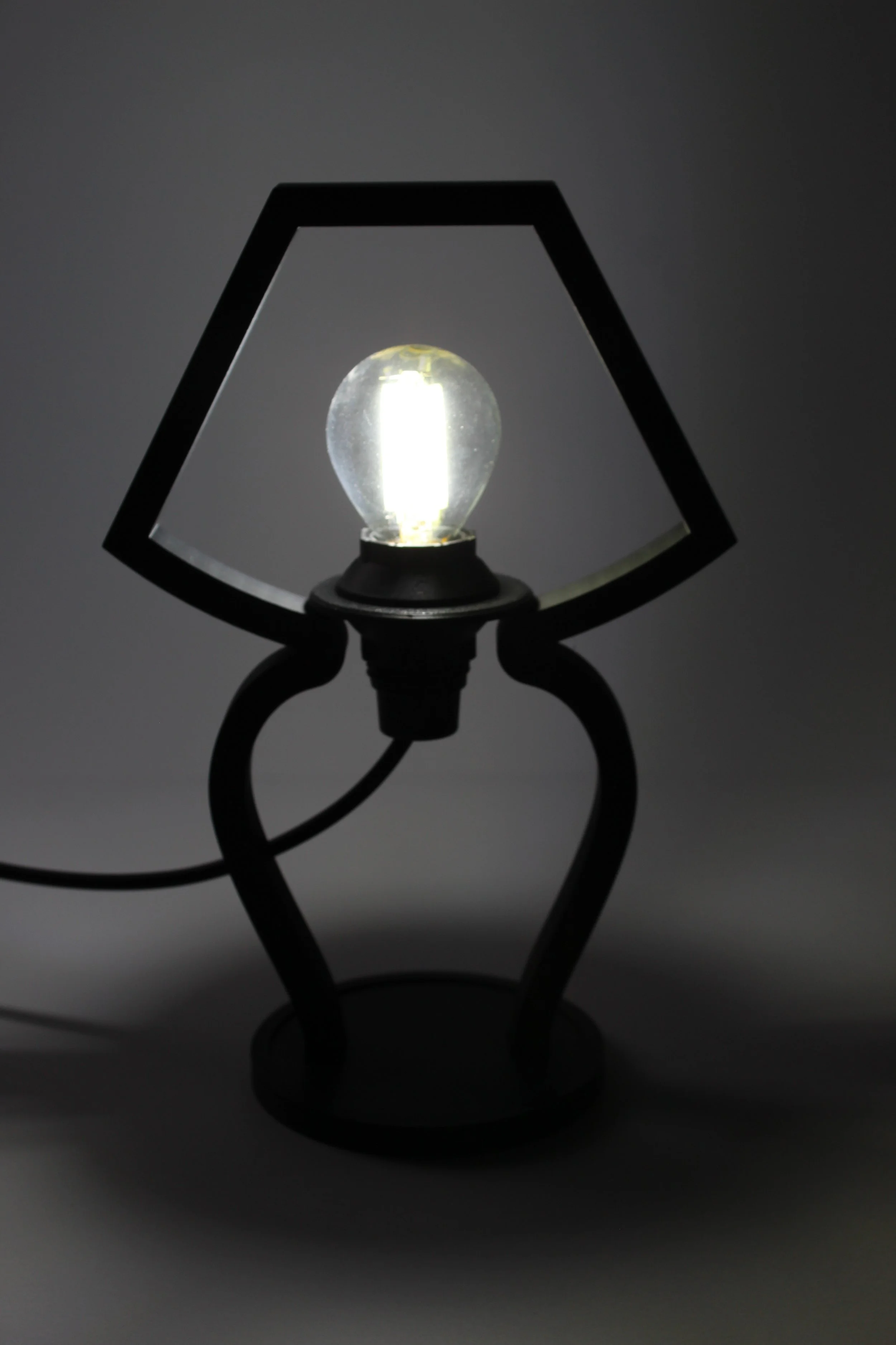 Lampada da tavolo con paralume a forma quadrata, lampadina accesa, sfondo grigio scuro.