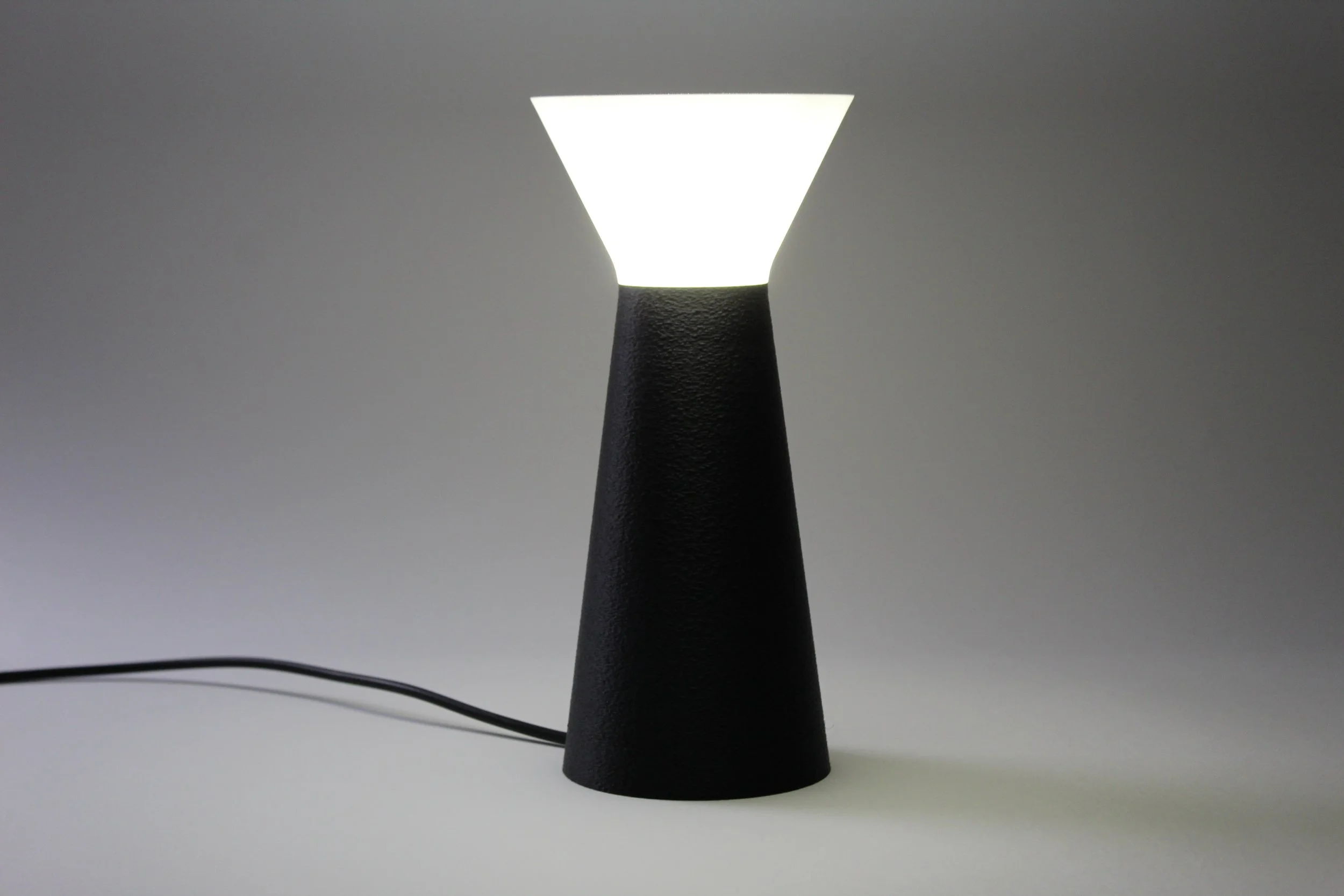 Lampada da tavolo con base nera e diffusore bianco a forma di cono rovesciato, accesa e posta su una superficie bianca