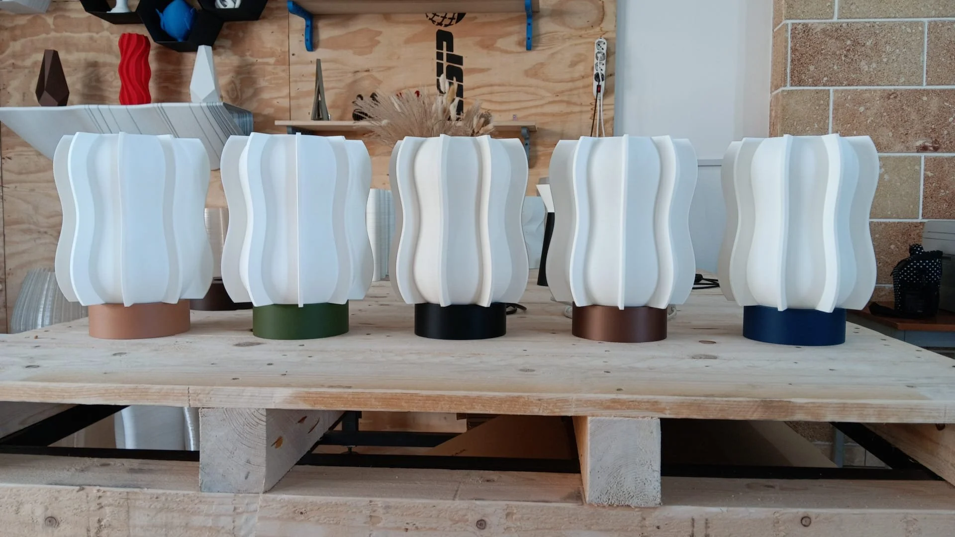 Cinque lampade da tavolo di design su un tavolo di legno, con basi di diversi colori e paralumi bianchi con forma ondulata.