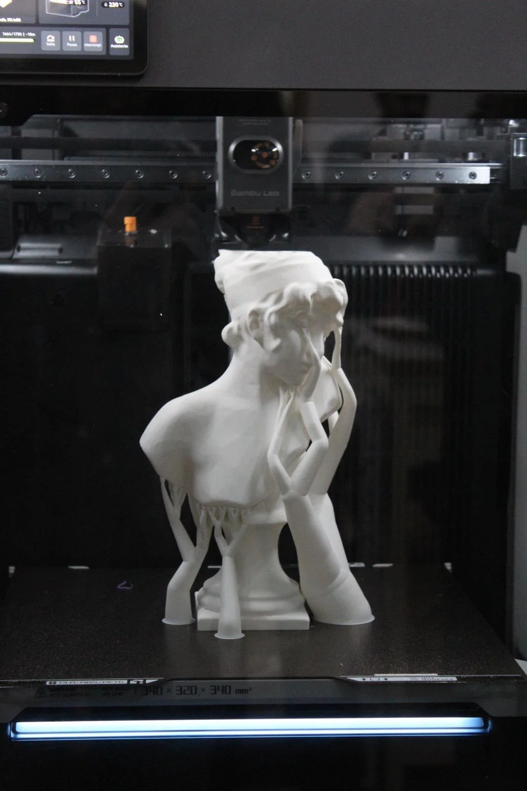 Statua di un volto femminile con capelli ricci, sovrapposta a un busto umano, in fase di stampa in una stampante 3D.