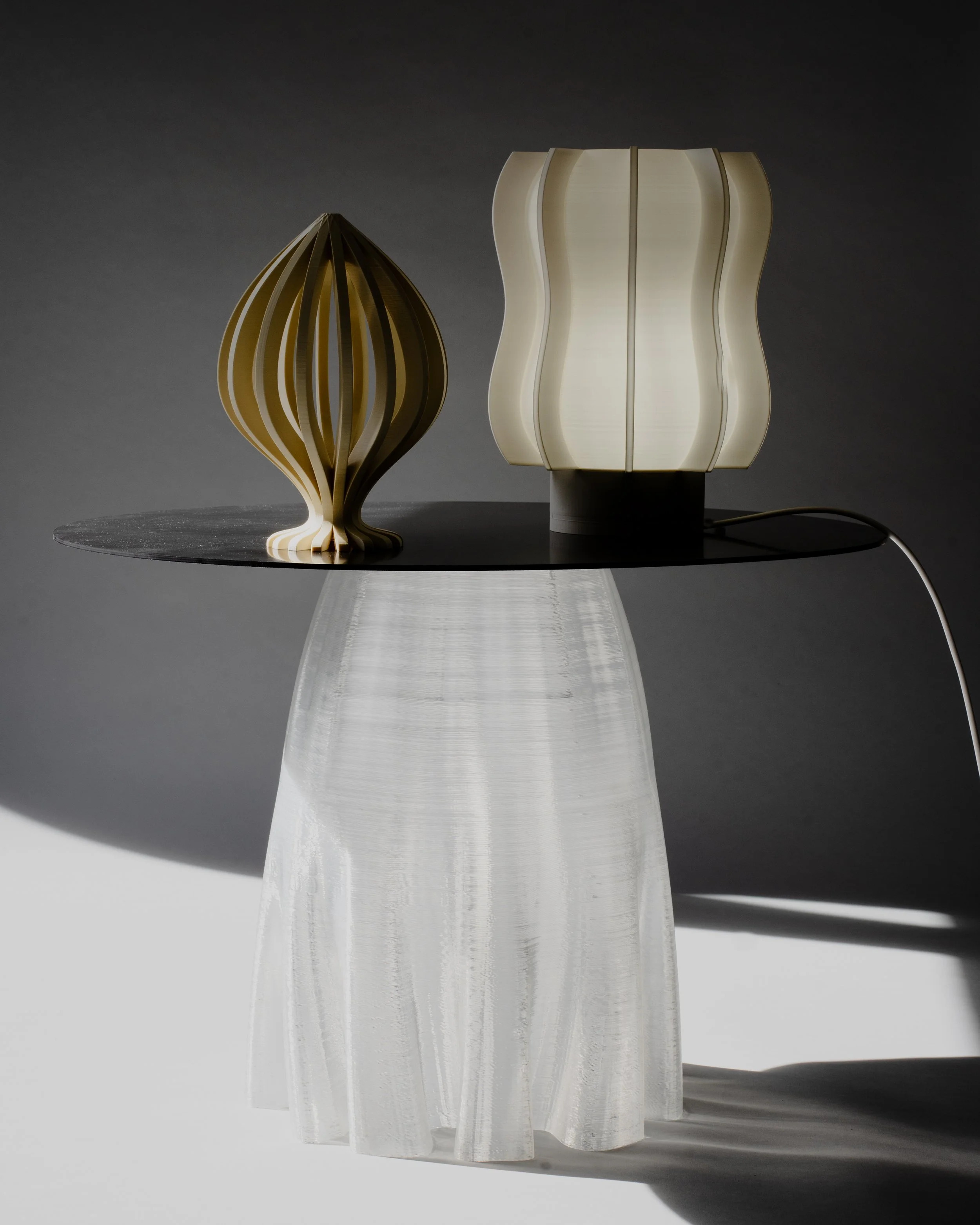 Due lampade moderne con design curvato e geometricamente strutturato, poste su un tavolo nero. La lampada a sinistra è in legno, mentre quella a destra è in materiale simile al plastica opaca.
