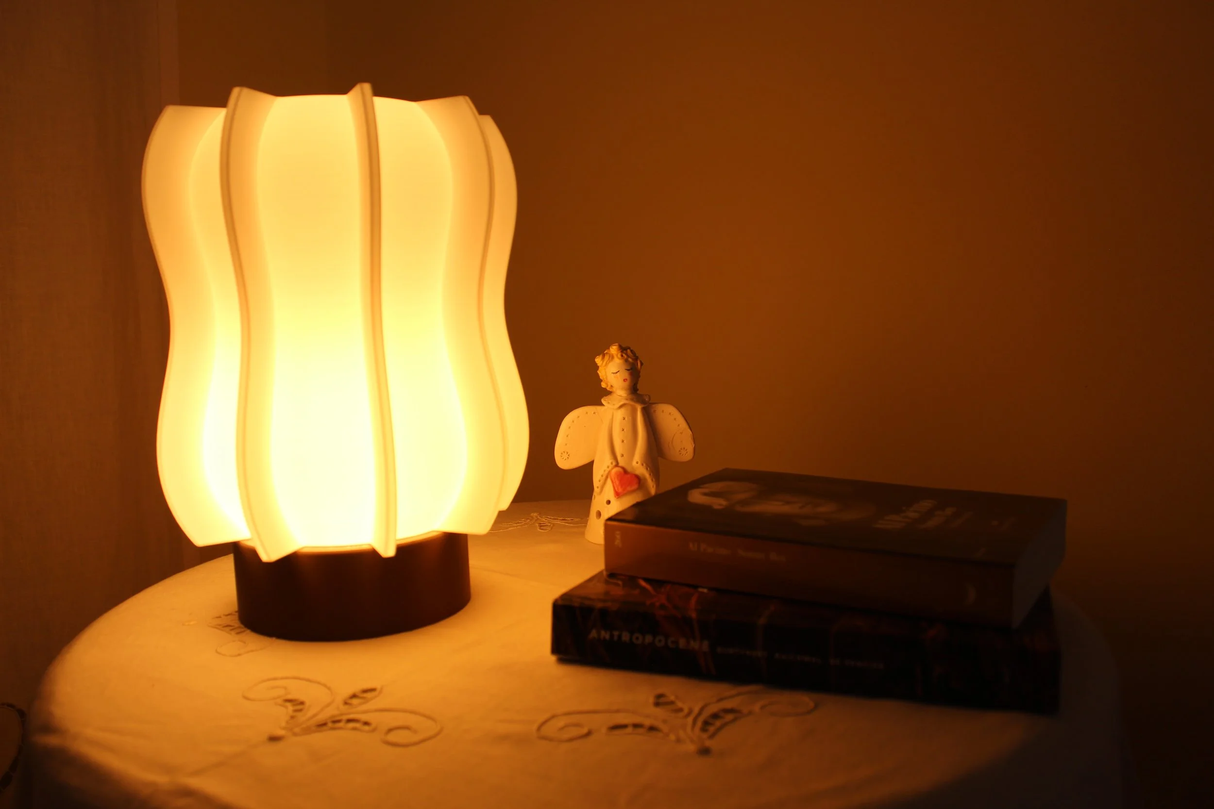 Lampada da tavolo accesa, statuetta di un angelo e due libri su un tavolo. Atmosfera calda e intima.