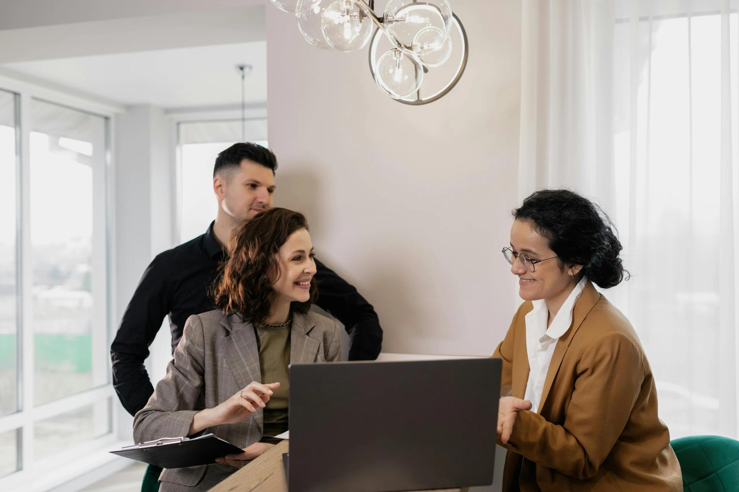 Four people having a meeting in an office, with a woman in glasses and a tan blazer talking to a woman with curly hair and a plaid blazer, while a man with dark hair looks on.