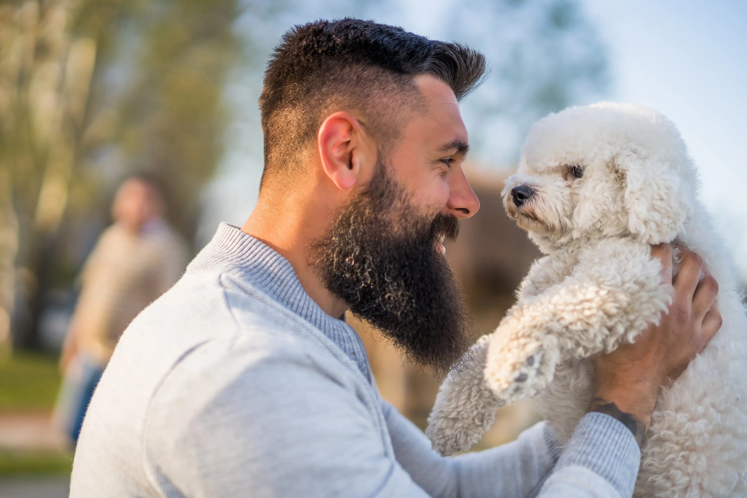 A man with a beard and short dark hair is holding a fluffy white dog up close to his face outdoors.
