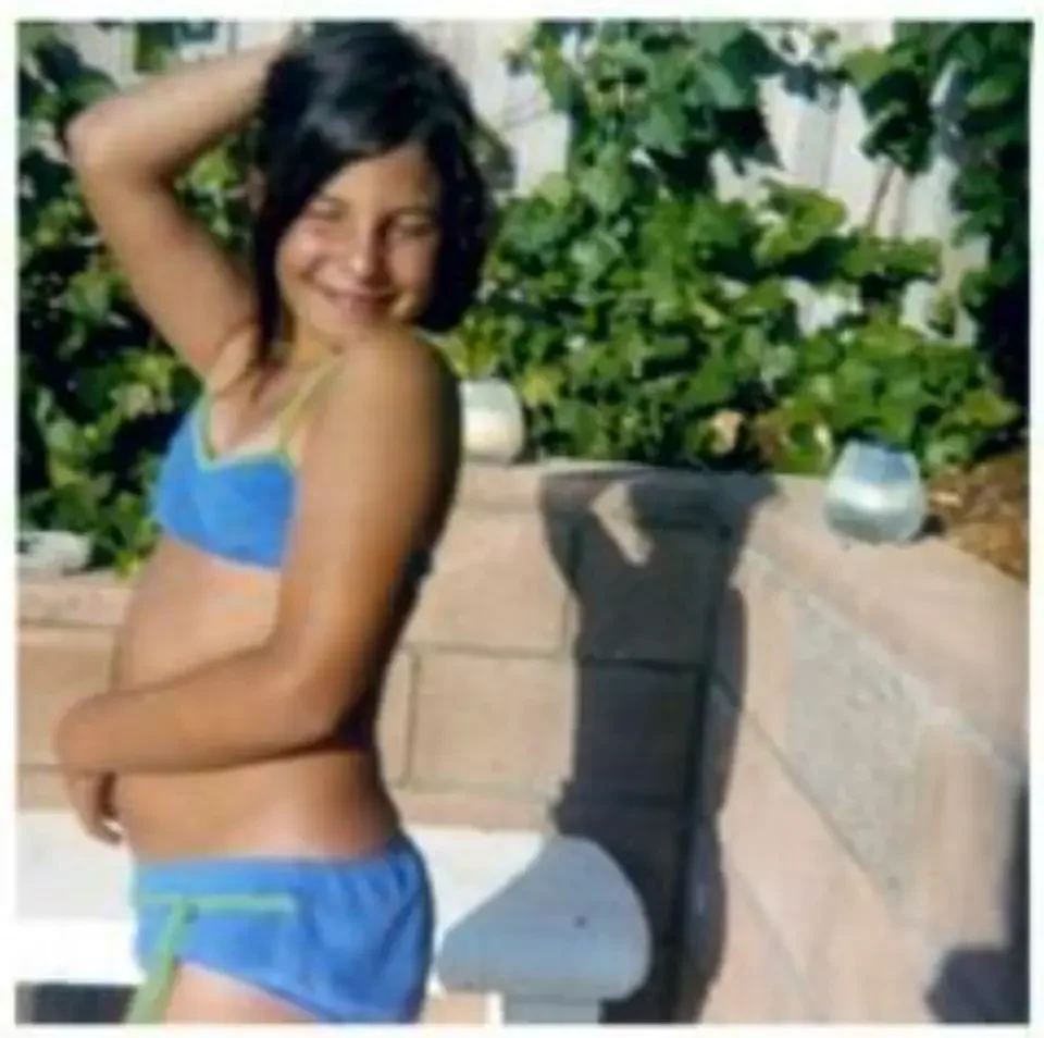 A young girl wearing a blue and yellow two-piece swimsuit, smiling with her eyes closed, standing outdoors in front of greenery and a white railing.