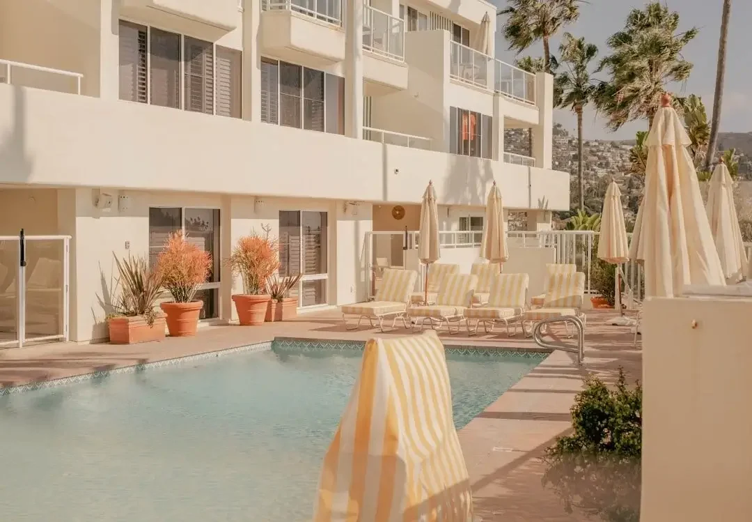 Hotel pool area with lounge chairs and umbrellas, potted plants, and a multi-story building with balconies in the background.