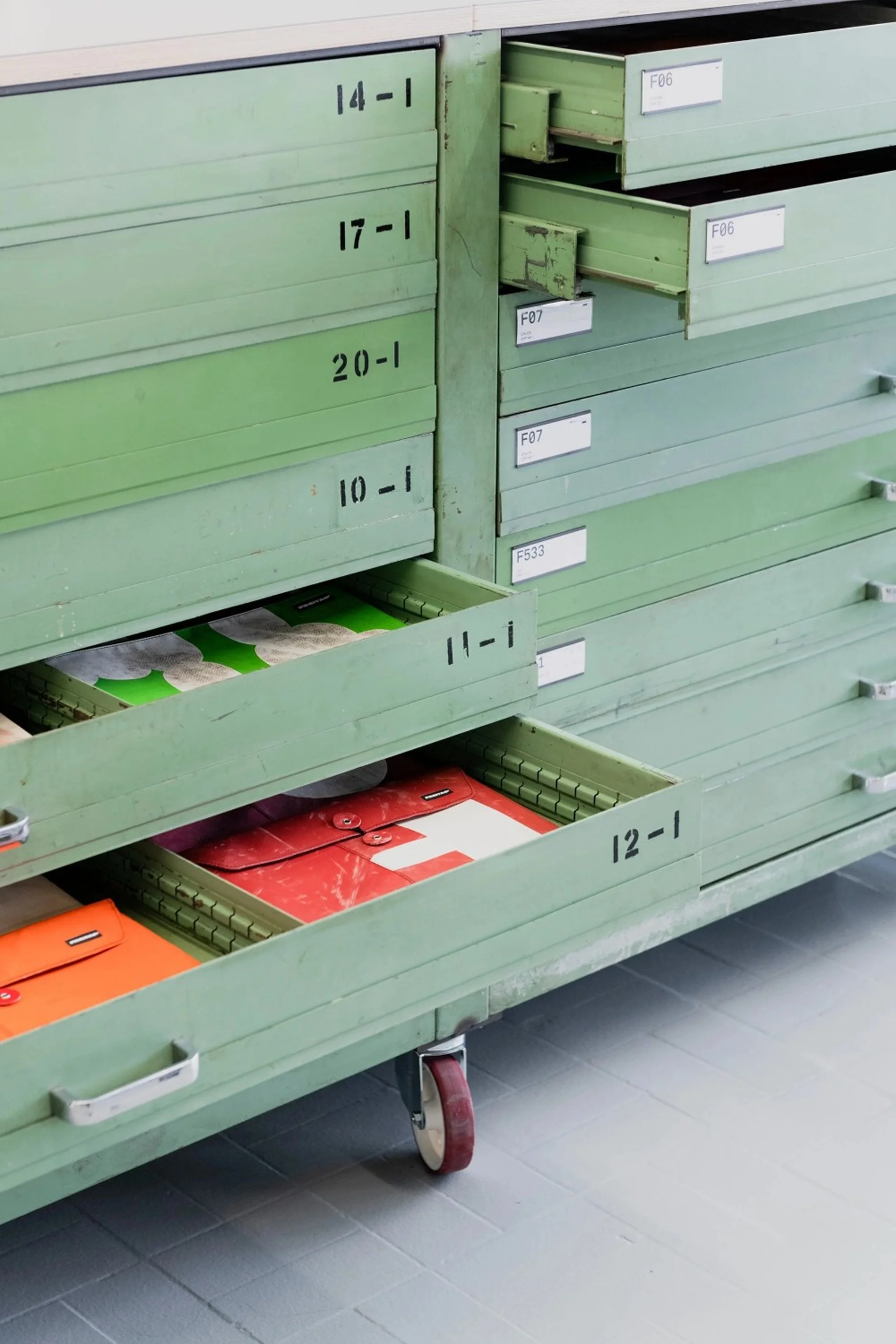 Flat files housed in folders in green drawers with labels, some partially open showing various folders and documents inside, on gray tiled floor.