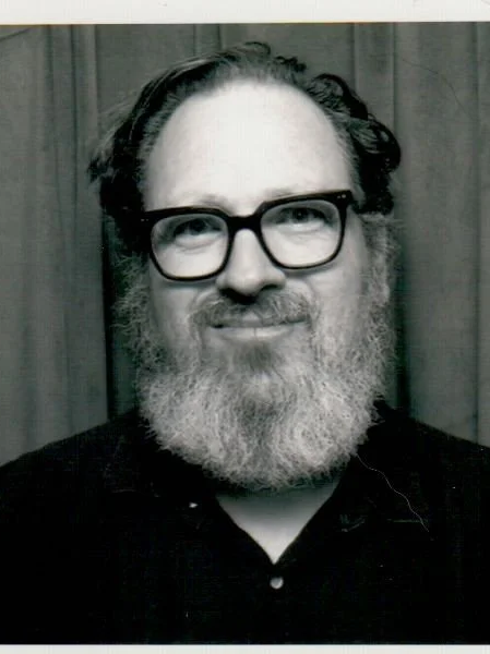 A man with glasses and a full beard, smiling, standing in front of curtains.