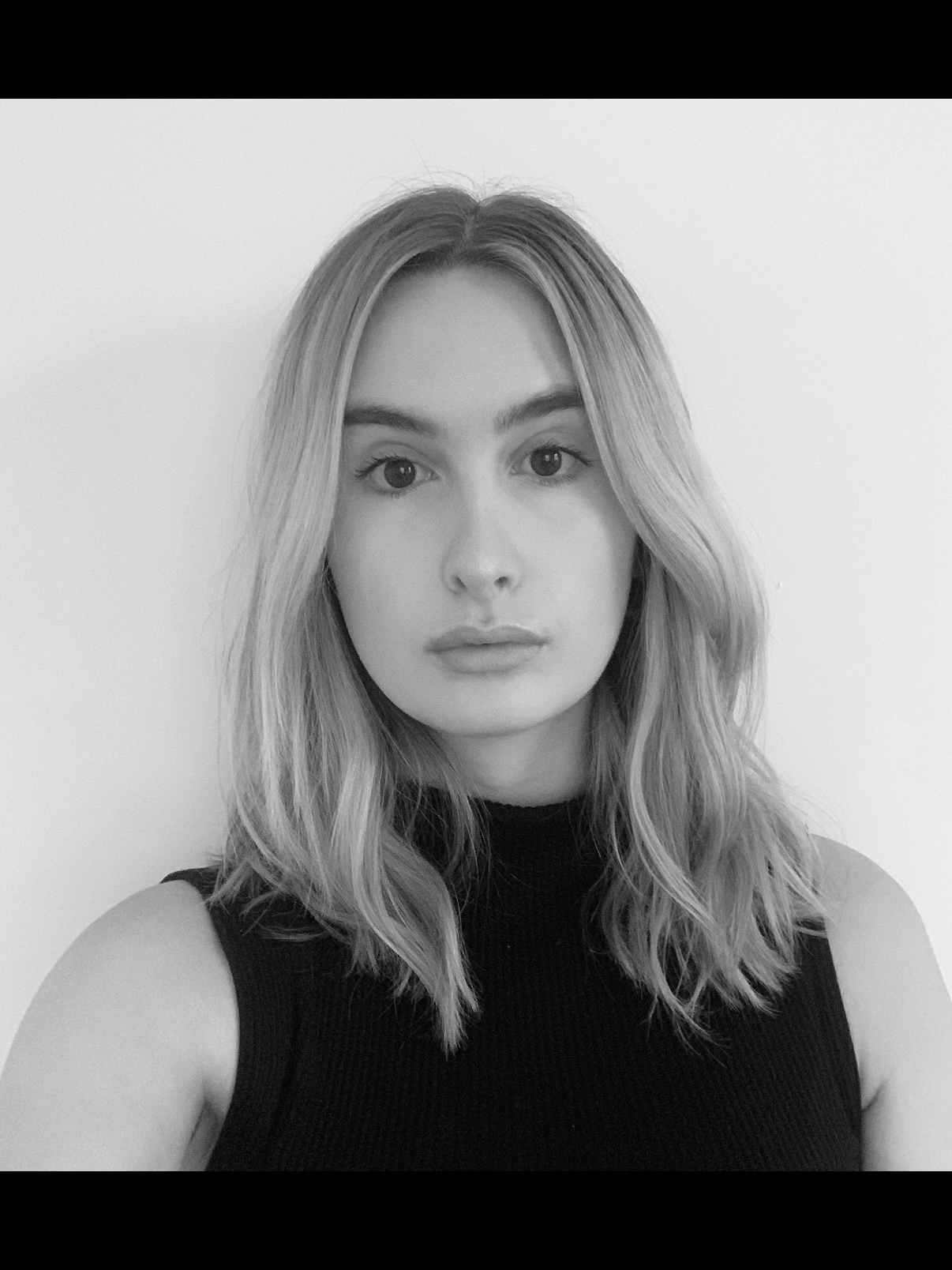 A black and white portrait of a young woman with shoulder-length wavy hair, looking directly at the camera, wearing a sleeveless top against a plain background.