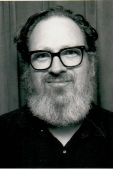 A black and white photo of a man with a beard, glasses, and curly hair, smiling at the camera.