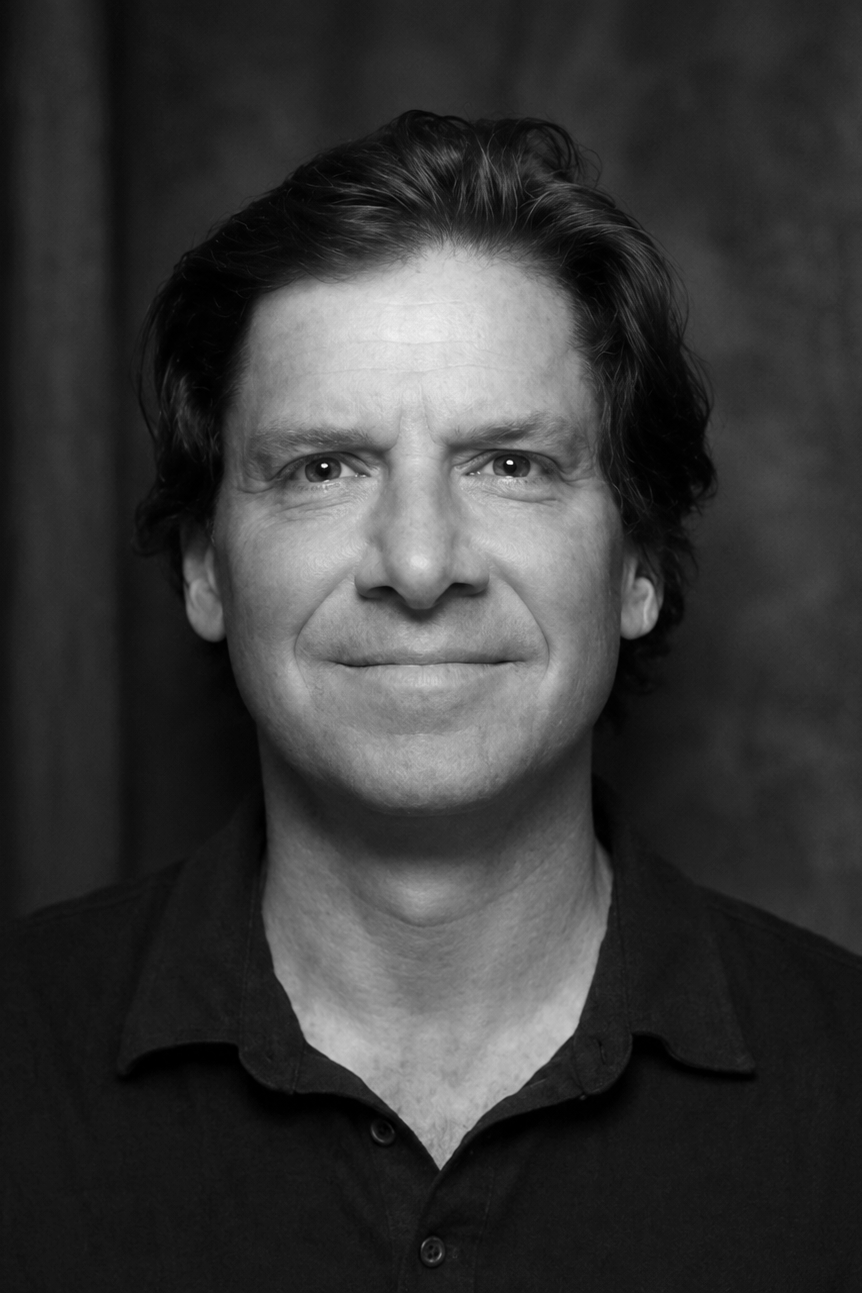 Black and white portrait of a man with dark, wavy hair, wearing a dark collared shirt, smiling slightly, against a dark background.