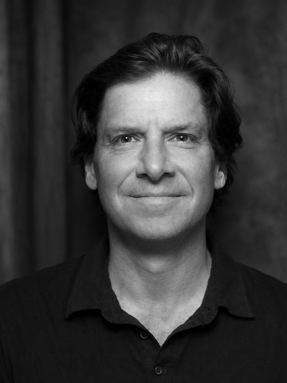 A black and white portrait of a man with medium-length wavy hair, wearing a collared shirt, smiling softly, with a textured background.