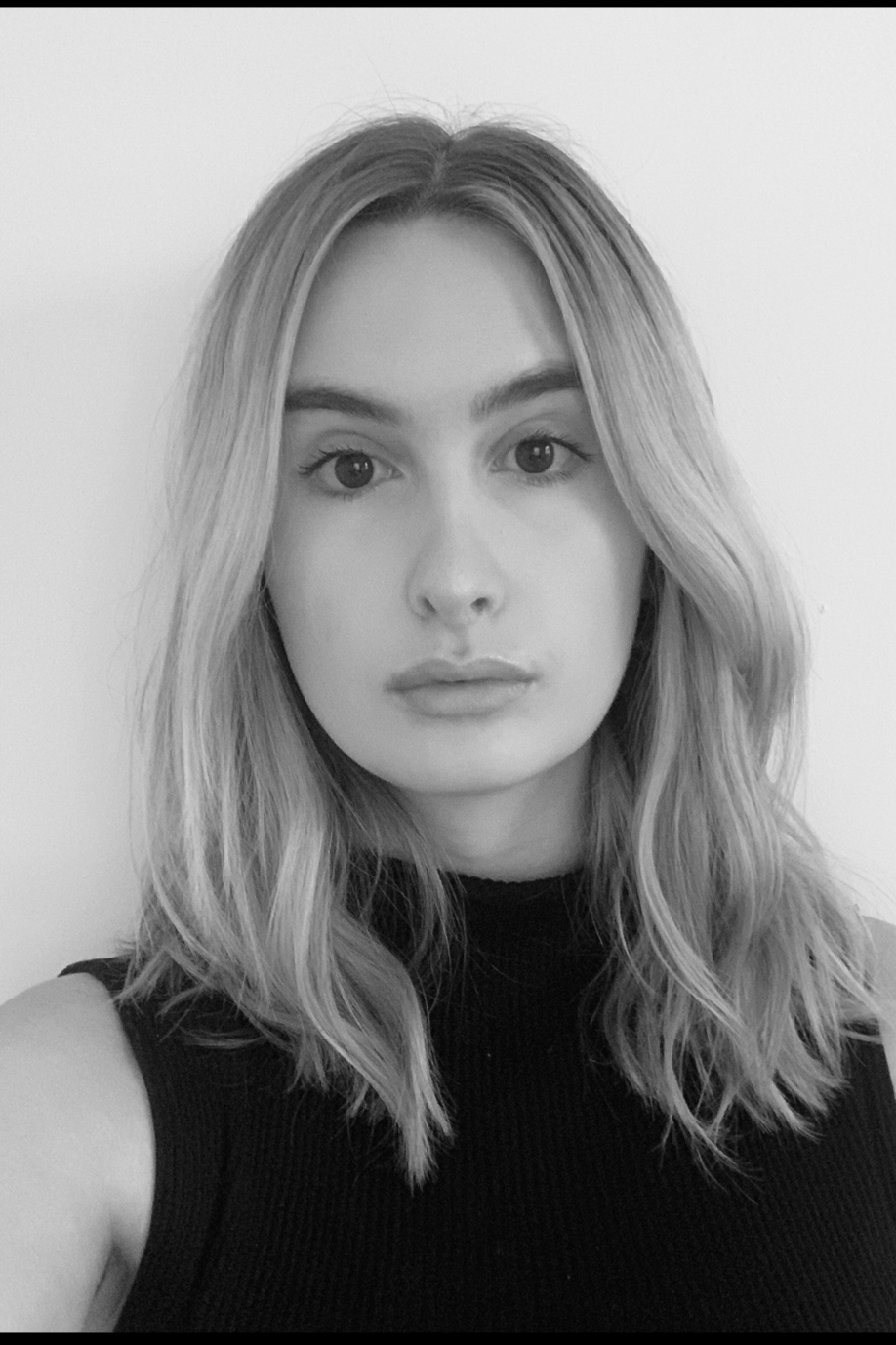 Black and white portrait of a young woman with shoulder-length wavy hair, wearing a sleeveless black top, looking directly at the camera with a neutral expression.