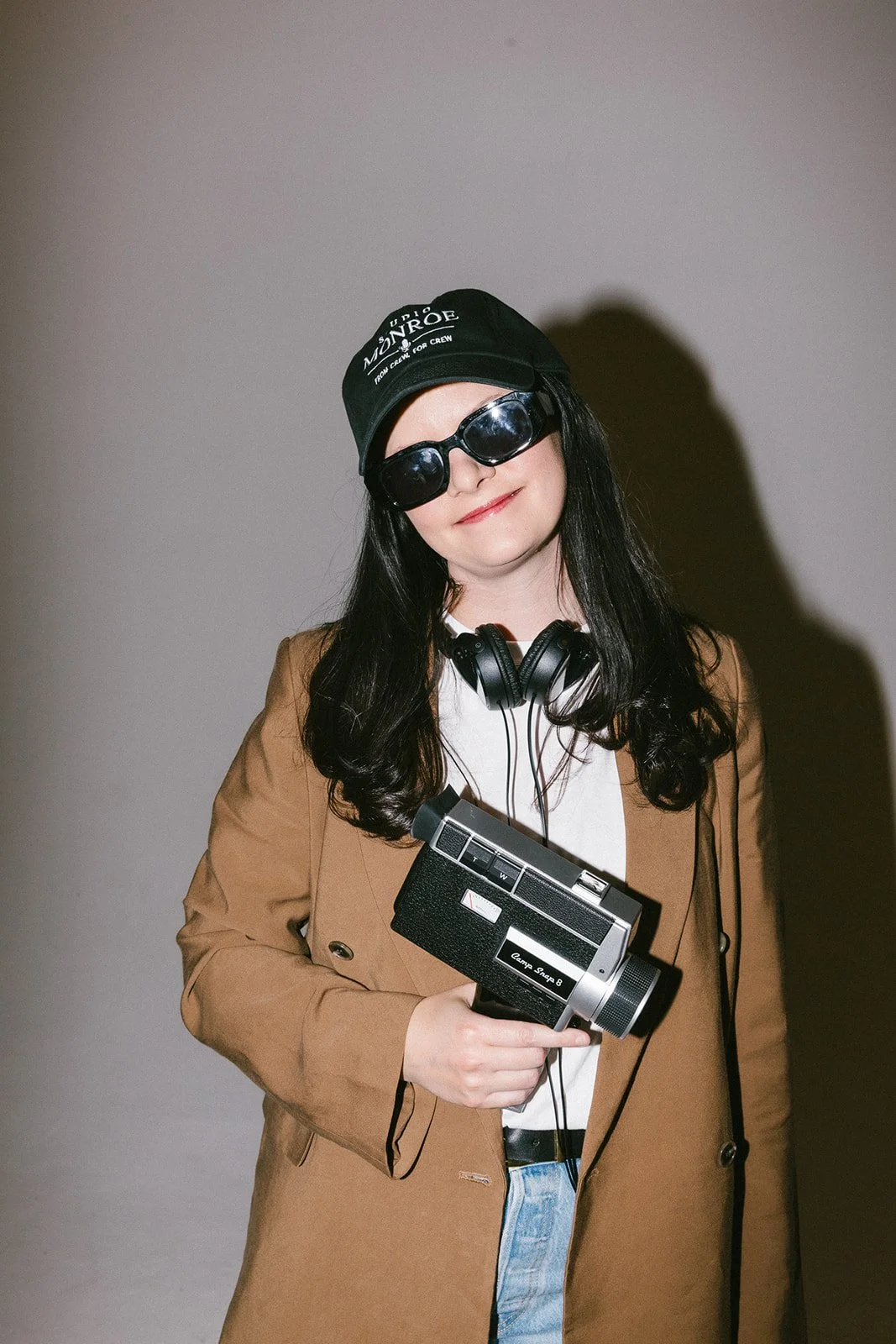 Woman with long black hair wearing a tan blazer, white shirt, black sunglasses, black cap, and headphones holding a vintage camcorder against a plain wall.