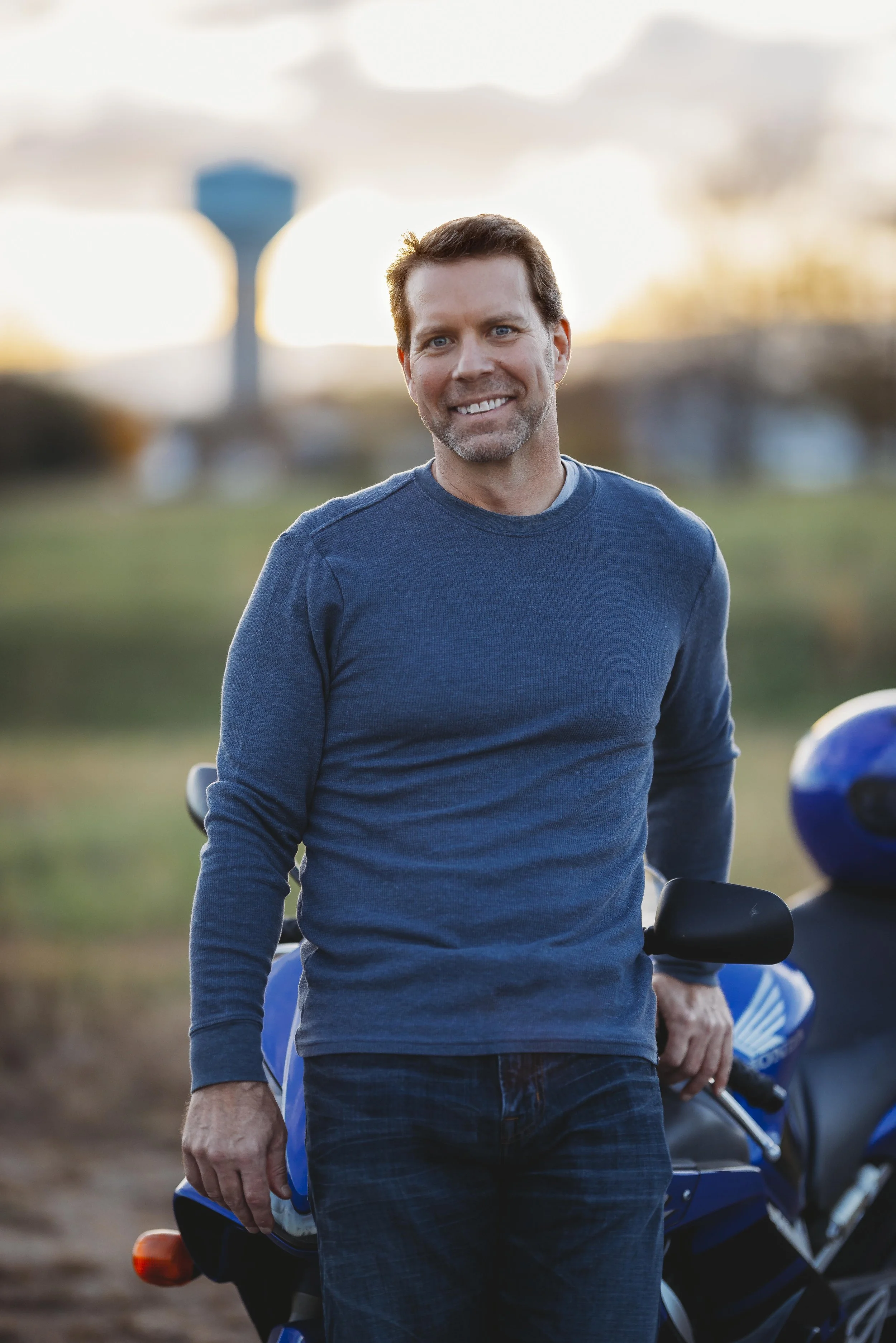 A middle-aged man with brown hair and a beard smiling outdoors, wearing a blue long-sleeve shirt, with a motorcycle and another person wearing a helmet in the background.