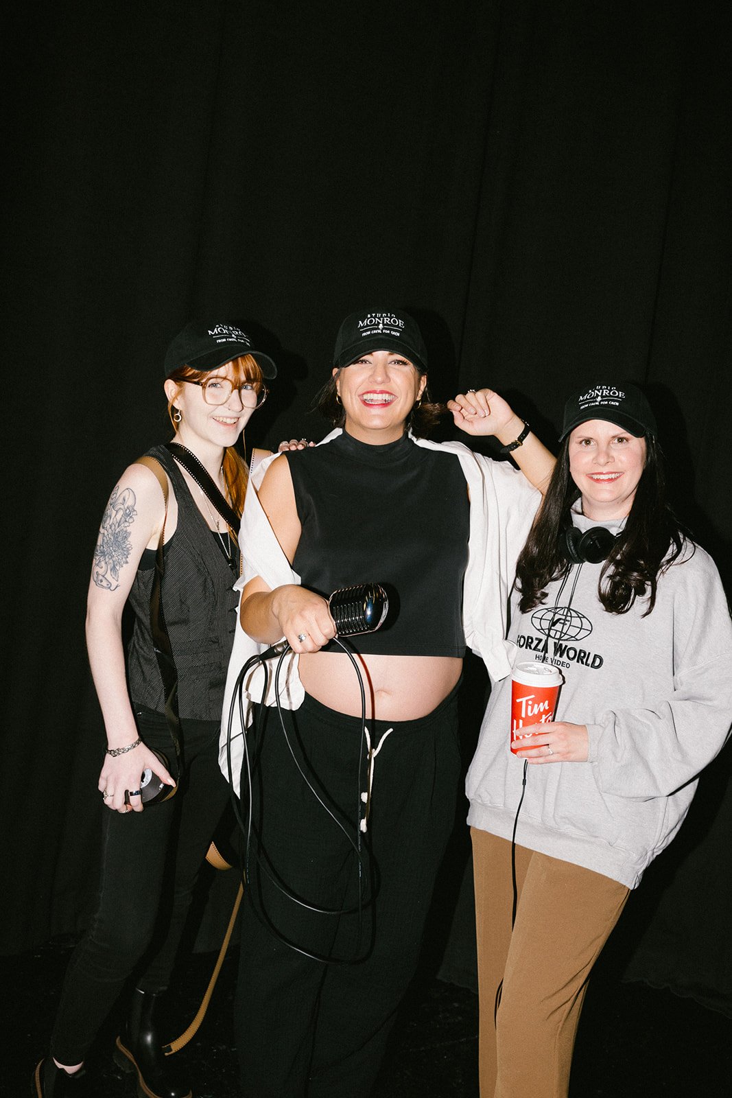 Three women posing together, wearing black hats with white writing, one woman in the middle holding a microphone, all smiling, against a black background.