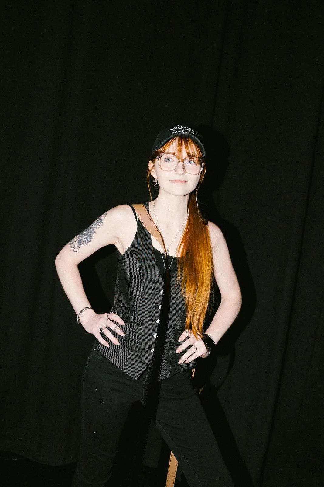 A woman with long red hair, glasses, and tattoos standing against a black background, wearing a black vest, black pants, and a black baseball cap.