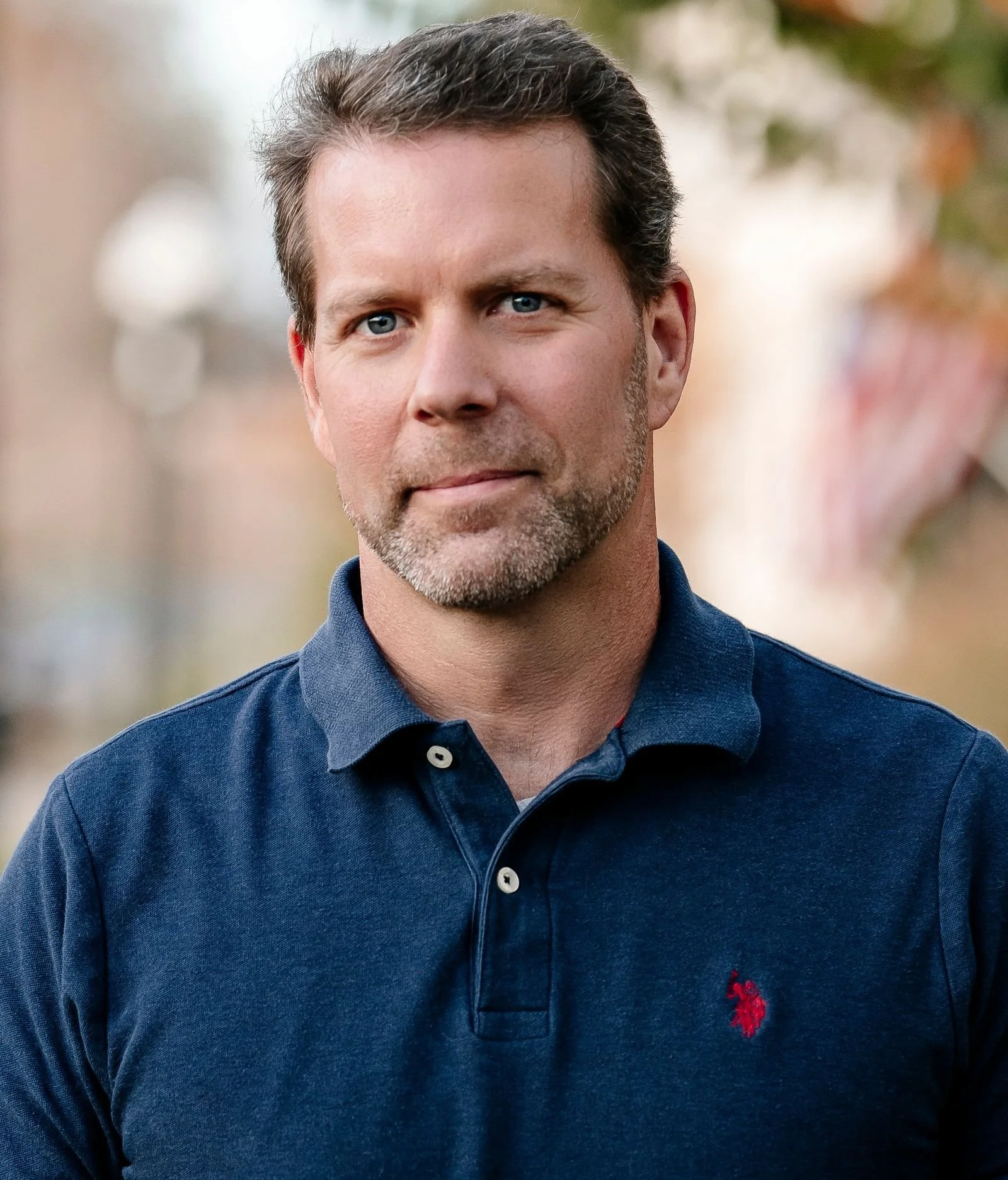 Close-up of a middle-aged man with blue eyes, short brown hair, and a light beard, wearing a navy blue polo shirt with a red Ralph Lauren logo.