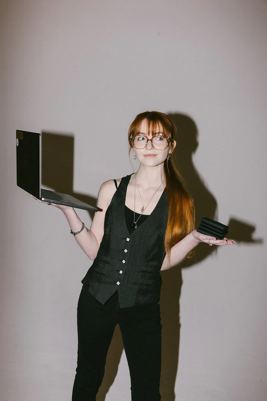 A woman with red hair and glasses is holding a laptop in her right hand and a black object in her left hand, standing against a plain wall.